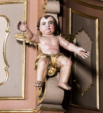 Zdjęcie nr 2: Przyścienna, wisząca ambona składa się z kosza, zaplecka i baldachimu. Wnętrze ambony dostępne jest przez drzwiczki umieszczone w polu zaplecka za pomocą schodów prowadzących z zakrystii. Kosz wykonano na planie prostokąta o wypukłej dłuższej ściance i wklęsłych krótszych, ustawiony na spłaszczonym wałku. Jej spód zdobi podwieszenie w formie zbliżonej do stożka, ujętego czterema wolutami, zakończone elementem wolutowym. W narożach kosza znalazły się cztery pilastry o trzonach przechodzących w spływy wolutowe, dźwigających imposty oraz parapet w formie gzymsu. Na frontowych wolutach umieszczono siedzące putta. Ścianki zostały dekorowane prostokątnymi płycinami. Na ściance frontowej wyodrębniono płycinę o wklęsło wypukłym zarysie z umieszczonymi w polu dwiema uskrzydlonymi główkami anielskimi oraz ornamentem roślinnym. Zaplecek uzyskał formę stojącego prostokąta ujętego parą pilastrów z konsolami dźwigającymi imposty oraz profilowany gzyms. W polu zamontowano drzwiczki dekorowane płycinami z uskrzydlonymi główkami anielskimi. Baldachim powstał w formie podstawy na planie analogicznym względem kosza, obwiedziony profilowanym gzymsem z fragmentami przerwanego wolutowego przyczółka, na którym siedzą putta. Całość zwieńczono medalionem w otoku z obłoków ze srebrzoną gołębicą Ducha Świętego i trzema główkami anielskimi w glorii promienistej. Struktura została polichromowana w kolorze pudrowego różu z jasnofioletowymi detalami, o złoconych profilach i glorii.