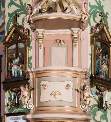 Zdjęcie nr 1: Przyścienna, wisząca ambona składa się z kosza, zaplecka i baldachimu. Wnętrze ambony dostępne jest przez drzwiczki umieszczone w polu zaplecka za pomocą schodów prowadzących z zakrystii. Kosz wykonano na planie prostokąta o wypukłej dłuższej ściance i wklęsłych krótszych, ustawiony na spłaszczonym wałku. Jej spód zdobi podwieszenie w formie zbliżonej do stożka, ujętego czterema wolutami, zakończone elementem wolutowym. W narożach kosza znalazły się cztery pilastry o trzonach przechodzących w spływy wolutowe, dźwigających imposty oraz parapet w formie gzymsu. Na frontowych wolutach umieszczono siedzące putta. Ścianki zostały dekorowane prostokątnymi płycinami. Na ściance frontowej wyodrębniono płycinę o wklęsło wypukłym zarysie z umieszczonymi w polu dwiema uskrzydlonymi główkami anielskimi oraz ornamentem roślinnym. Zaplecek uzyskał formę stojącego prostokąta ujętego parą pilastrów z konsolami dźwigającymi imposty oraz profilowany gzyms. W polu zamontowano drzwiczki dekorowane płycinami z uskrzydlonymi główkami anielskimi. Baldachim powstał w formie podstawy na planie analogicznym względem kosza, obwiedziony profilowanym gzymsem z fragmentami przerwanego wolutowego przyczółka, na którym siedzą putta. Całość zwieńczono medalionem w otoku z obłoków ze srebrzoną gołębicą Ducha Świętego i trzema główkami anielskimi w glorii promienistej. Struktura została polichromowana w kolorze pudrowego różu z jasnofioletowymi detalami, o złoconych profilach i glorii.