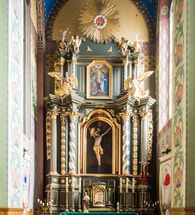 Zdjęcie nr 1: Nastawa ołtarza Ukrzyżowania uzyskała formę architektonicznego retabulum dwukondygnacyjnego, jednoosiowego ze zwieńczeniem w postaci promienistej glorii. Drewniany stipes ołtarzowy uzyskał formę prostopadłościanu podtrzymującego drewnianą mensę z marmurowym portatylem. Na mensie na osi ustawiono architektoniczne tabernakulum w formie drewnianej skrzynki na cokole, na planie trapezu o wypukłej ściance przedniej i wklęsłych ramionach. Ścianka przednia ujęta została parą zdwojonych pilastrów podtrzymujących imposty, których trzon przyjmuje formę wolut. Na ściance przedniej usytuowana została nisza obrotowa w formie stojącego prostokąta zamkniętego łukiem półkolistym nadwieszonym z uskokiem w karbowanej ramie. Widoczne pilastry dźwigają pełne belkowanie, zaś nad polem głównym wyłącznie gzyms. Drzwiczki tabernakulum ozdobiono płaskorzeźbionym przedstawieniem baranka leżącego na księdze o siedmiu pieczęciach w glorii promienistej. Retabulum ustawiono na trójstrefowym cokole. W dwóch pierwszych strefach znalazły się dwie pary wyodrębnionych prostopadłościanów stanowiących cokoły dla kolumn. Nastawę flankują dwie pary kolumn o kapitelach korynckich, spośród których wewnętrza ma skręcone trzony, a zewnętrzna kanelowane. Kolumny dźwigają pełne belkowanie wyłamane na ich osi, pod kątem, zaś nad polem środkowym wyłącznie gzyms. Niemal w całości pole główne wypełnia wnęka w formie stojącego prostokąta zamkniętego łukiem odcinkowym, nadwieszonym z uskokiem, ujęta profilowaną ramą. Wewnątrz widoczna jest mniejsza w formie stojącego prostokąta zamkniętego łukiem odcinkowym z figurą Chrystusa Ukrzyżowanego. Druga kondygnacja ustawiona jest także na cokole. Flankują ją dwie pary kolumn o kapitelach korynckich, spośród których wewnętrza ma skręcone trzony, zewnętrzna kanelowane. Kolumny dźwigają pełne belkowanie wyłamane na ich osi, pod kątem oraz fragmenty półkolistego przyczółka. W polu umieszczono obraz Zwiastowania w formie stojącego prostokąta zamkniętego trójbocznie, ujętego ramą zdobioną wolim okiem. Całość zakończona szczytem ujętym wolutami z medalionem z monogramem "IHS" w otoku z obłoków w glorii promienistej. Cokół dolny zdobiony został płycinami i rozetami. Pomiędzy kolumnami usytuowano zwisy kwiatowe na wstędze. Uszy ołtarza utworzone zostały z akantu. Fryzy występujące w retabulum dekorowany są złoconymi rozetami, zaś przyłucza ozdobiono ornamentem roślinnym. Drugą kondygnację flankują uszy utworzone z akantu i wstęgi. Na belkowaniu umieszczono siedzące figury aniołów, a na drugiej kondygnacji putta i wazony. Struktura ołtarza została polichromowana w kolorze szaro-zielonym z elementami szaro-brązowymi ze złoconymi profilami, ramami i ornamentyką, natomiast kręcone trzony posrebrzono.