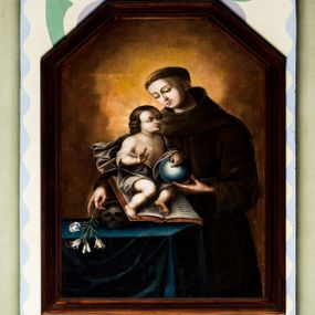 Zdjęcie nr 1: Obraz uzyskał kształt stojącego prostokąta zamkniętego trójbocznie. W centrum kompozycji ukazano św. Antoniego ujętego w trzech czwartych w lewo na tle poświaty rozjaśniającej brązowe tło. Święty z pochyloną głową, prawym ramieniem obejmuje Dzieciątko, które siedzi na otwartej księdze wspartej o czaszkę leżącą na stole. W tej samej dłoni Antoni trzyma lilię, lewą zaś ręką podtrzymuje błękitny glob z krzyżykiem, wsparty o kolana małego Jezusa. Święty ma owalną, młodzieńczą twarz o jasnej karnacji. Charakteryzują ją duże, półprzymknięte oczy, wydany nos i usta o pełnych wargach. Głowę otaczają krótkie włosy z wyciętą tonsurą, odsłaniające lewe ucho. Antoni ubrany jest w brązowy habit franciszkański. Mały Jezus ujęty został w trzech czwartych w prawo. Z podkurczonymi nogami siedzi na otwartej księdze, prawą ręką udzielając błogosławieństwa, lewą podtrzymując glob leżący na jego kolanach. Jezus ma pucułowatą twarz o jasnej karnacji, ciemne oczy, wydatny nos i usta. Jego głowę otaczają brązowe włosy zakończone lokami opadającymi na plecy. Zbawiciel ma nagi tors, jedynie, rozwiana, szarobrązowa tkanina przykrywa jego biodra i część ud. Wokół głowy widoczne są świetliste promienie. Stół na którym leży czaszka i księga nakryty jest ciemnoniebieską tkaniną.  
