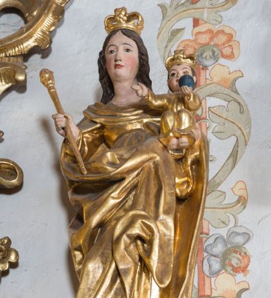 Zdjęcie nr 1: Figura ustawiona na niskiej, nieregularnej podstawie oraz na cokole ujętym spływami wolutowymi z monogramem maryjnym na froncie. Matka Boska ukazana frontalnie, przechylona na prawo, lewą ręką podtrzymuje Dzieciątko, w prawej trzyma berło. Twarz ma owalną, o migdałowatych oczach, długim nosie i wąskich ustach, okoloną długimi, brązowymi, falowanymi włosami. Ubrana jest w suknię oraz obfity, silnie drapowany płaszcz zarzucony na ramiona i zawinięty z przodu postaci. Dzieciątko w pozycji siedzącej, ze skulonymi nogami, zwrócone frontalnie, lewą rękę z jabłkiem królewskim wyciąga przed siebie, prawą w geście błogosławieństwa wyciąga w bok. Twarz ma kwadratową, o pełnych policzkach, dużych oczach, małym nosie i ustach, okoloną krótkimi, brązowymi włosami. Ubrane jest w tunikę. Na głowach obu postaci korony zamknięte. Figury polichromowane naturalistycznie w partiach ciała, szaty i atrybuty złocone. 