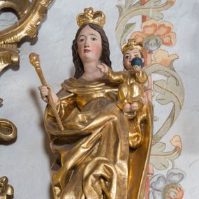 Zdjęcie nr 1: Figura ustawiona na niskiej, nieregularnej podstawie oraz na cokole ujętym spływami wolutowymi z monogramem maryjnym na froncie. Matka Boska ukazana frontalnie, przechylona na prawo, lewą ręką podtrzymuje Dzieciątko, w prawej trzyma berło. Twarz ma owalną, o migdałowatych oczach, długim nosie i wąskich ustach, okoloną długimi, brązowymi, falowanymi włosami. Ubrana jest w suknię oraz obfity, silnie drapowany płaszcz zarzucony na ramiona i zawinięty z przodu postaci. Dzieciątko w pozycji siedzącej, ze skulonymi nogami, zwrócone frontalnie, lewą rękę z jabłkiem królewskim wyciąga przed siebie, prawą w geście błogosławieństwa wyciąga w bok. Twarz ma kwadratową, o pełnych policzkach, dużych oczach, małym nosie i ustach, okoloną krótkimi, brązowymi włosami. Ubrane jest w tunikę. Na głowach obu postaci korony zamknięte. Figury polichromowane naturalistycznie w partiach ciała, szaty i atrybuty złocone. 