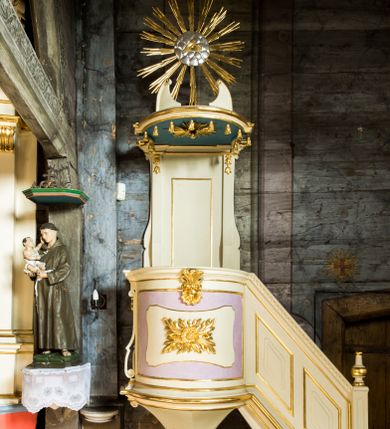 Zdjęcie nr 1: Przyścienna, wisząca ambona składa się z kosza, zaplecka i baldachimu oraz prowadzący na nią jednobiegowych schodów z balustradą. Kosz został wykonany na planie prostokąta z półkoliście wybrzuszoną ścianką frontową oraz wklęsłą boczną, z podwieszeniem w formie połowy stożka o wklęsłych ścianach, zakończonego wolutą. Ustawiono go na profilowanym cokole. Ściankę boczną ujęto parą pilastrów o trzonach przechodzących w spływy wolutowe, dekorowane kampanulami. Zarówno ją, jak i frontową wypełniono płycinami z wydzielonymi w centrum fantazyjnymi polami, od frontu dekorowaną rozetą oraz stylizowanym anthemionem z muszlą. Kosz zwieńczono parapetem w formie gzymsu. Balustrada schodów powstała jako pełna, z profilowanym cokołem, polem wypełnionym romboidalnymi płycinami z wydzielonymi fantazyjnymi polami wewnątrz, zamknięta profilowanym gzymsem z kostkowaniem. Balustradę zamyka filarek na cokole, o trzonie dekorowanym płycinami, na którym stoi wazon. Zaplecek ujęty został wąskimi pilastrami podtrzymującymi profilowany gzyms, o trzonach dekorowanych kampanulami. Pole udekorowano płyciną. Baldachim wykonano w formie podestu na planie prostokąta z półkoliście wybrzuszoną ścianką frontową oraz wklęsłymi bocznymi, z uszakami. Całoś okala profilowany gzyms z podwieszonymi chwostami. Na baldachimie usytuowano fragmenty przerwanego falistego przyczółka oraz oko Opatrzności Bożej na tle srebrnych obłoków w złotej glorii promienistej. Na podniebieniu baldachimu zawieszono rzeźbioną gołębicę Ducha Świętego w glorii. Struktura została polichromowana na biało z jasnofioletowymi polami, jasnoniebieskim podniebieniem baldachimu, ze złoconymi profilami i ornamentami oraz srebrzonymi detalami.