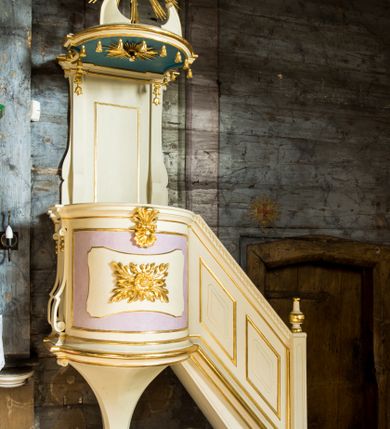 Zdjęcie nr 2: Przyścienna, wisząca ambona składa się z kosza, zaplecka i baldachimu oraz prowadzący na nią jednobiegowych schodów z balustradą. Kosz został wykonany na planie prostokąta z półkoliście wybrzuszoną ścianką frontową oraz wklęsłą boczną, z podwieszeniem w formie połowy stożka o wklęsłych ścianach, zakończonego wolutą. Ustawiono go na profilowanym cokole. Ściankę boczną ujęto parą pilastrów o trzonach przechodzących w spływy wolutowe, dekorowane kampanulami. Zarówno ją, jak i frontową wypełniono płycinami z wydzielonymi w centrum fantazyjnymi polami, od frontu dekorowaną rozetą oraz stylizowanym anthemionem z muszlą. Kosz zwieńczono parapetem w formie gzymsu. Balustrada schodów powstała jako pełna, z profilowanym cokołem, polem wypełnionym romboidalnymi płycinami z wydzielonymi fantazyjnymi polami wewnątrz, zamknięta profilowanym gzymsem z kostkowaniem. Balustradę zamyka filarek na cokole, o trzonie dekorowanym płycinami, na którym stoi wazon. Zaplecek ujęty został wąskimi pilastrami podtrzymującymi profilowany gzyms, o trzonach dekorowanych kampanulami. Pole udekorowano płyciną. Baldachim wykonano w formie podestu na planie prostokąta z półkoliście wybrzuszoną ścianką frontową oraz wklęsłymi bocznymi, z uszakami. Całoś okala profilowany gzyms z podwieszonymi chwostami. Na baldachimie usytuowano fragmenty przerwanego falistego przyczółka oraz oko Opatrzności Bożej na tle srebrnych obłoków w złotej glorii promienistej. Na podniebieniu baldachimu zawieszono rzeźbioną gołębicę Ducha Świętego w glorii. Struktura została polichromowana na biało z jasnofioletowymi polami, jasnoniebieskim podniebieniem baldachimu, ze złoconymi profilami i ornamentami oraz srebrzonymi detalami.