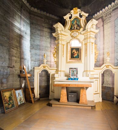Zdjęcie nr 2: Jednokondygnacyjna i jednoosiowa nastawa ołtarza głównego uzyskała formę architektoniczną ze zwieńczeniem, ustawioną na wysokim cokole, flankowaną przez dwie bramki. Drewniany stipes ołtarzowy uzyskał kształt prostopadłościanu podtrzymującego drewnianą mensę z portatylem. Nastawa została ujęta parą opilastrowanych filarów, ustawionych pod kątem, zwróconych na zewnątrz, dźwigających pełne belkowanie przerwane nad polem środkowym oraz fragmenty przerwanego odwróconego przyczółka. Trzony pilastrów dekorowane zostały rocaille’owymi zwisami. W polu głównym znajduje się prześwit w formie stojącego prostokąta zamkniętego łukiem półkolistym nadwieszonym z umieszczonym wtórnie mniejszym obrazem Matki Boskiej z Dzieciątkiem. Nad nim usytuowano owalny kartusz ujęty ornamentem roślinnym z monogramem Marii w polu. Kondygnację flankują uszaki utworzone z ażurowych elementów rocaille. Zwieńczenie uzyskało formę trapezu o wklęsłych bokach zakończonego trzema wypustami zamkniętymi fragmentami gzymsu przechodzącego w woluty. W polu umieszczono obraz św. Wojciecha w profilowanej ramie w formie zbliżonej do trapezu o wklęsłych bokach i ściętych dolnych narożach, zamkniętego łukiem falistym. Zwieńczenie ujęte zostało liśćmi palmowymi, a zakończone muszlą i anthemionem. Bramki zostały zamknięte profilowanym gzymsem wyłamującym się po środku trójbocznie ze stojącymi na nim wazami płomienistymi. Przejścia utworzono w formie stojącego prostokąta zamkniętego łukiem półkolistym nadwieszonym. Obwiedziono je profilowaną ramą, zakończoną rozetami. Struktura nastawy została polichromowana w kolorze kości słoniowej, ze złoconymi profilami i ornamentami.