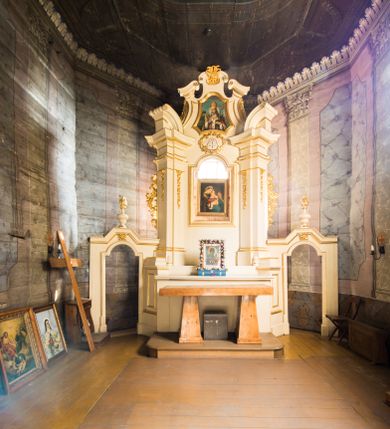 Zdjęcie nr 1: Jednokondygnacyjna i jednoosiowa nastawa ołtarza głównego uzyskała formę architektoniczną ze zwieńczeniem, ustawioną na wysokim cokole, flankowaną przez dwie bramki. Drewniany stipes ołtarzowy uzyskał kształt prostopadłościanu podtrzymującego drewnianą mensę z portatylem. Nastawa została ujęta parą opilastrowanych filarów, ustawionych pod kątem, zwróconych na zewnątrz, dźwigających pełne belkowanie przerwane nad polem środkowym oraz fragmenty przerwanego odwróconego przyczółka. Trzony pilastrów dekorowane zostały rocaille’owymi zwisami. W polu głównym znajduje się prześwit w formie stojącego prostokąta zamkniętego łukiem półkolistym nadwieszonym z umieszczonym wtórnie mniejszym obrazem Matki Boskiej z Dzieciątkiem. Nad nim usytuowano owalny kartusz ujęty ornamentem roślinnym z monogramem Marii w polu. Kondygnację flankują uszaki utworzone z ażurowych elementów rocaille. Zwieńczenie uzyskało formę trapezu o wklęsłych bokach zakończonego trzema wypustami zamkniętymi fragmentami gzymsu przechodzącego w woluty. W polu umieszczono obraz św. Wojciecha w profilowanej ramie w formie zbliżonej do trapezu o wklęsłych bokach i ściętych dolnych narożach, zamkniętego łukiem falistym. Zwieńczenie ujęte zostało liśćmi palmowymi, a zakończone muszlą i anthemionem. Bramki zostały zamknięte profilowanym gzymsem wyłamującym się po środku trójbocznie ze stojącymi na nim wazami płomienistymi. Przejścia utworzono w formie stojącego prostokąta zamkniętego łukiem półkolistym nadwieszonym. Obwiedziono je profilowaną ramą, zakończoną rozetami. Struktura nastawy została polichromowana w kolorze kości słoniowej, ze złoconymi profilami i ornamentami.