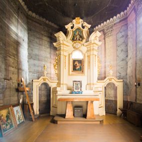 Zdjęcie nr 1: Jednokondygnacyjna i jednoosiowa nastawa ołtarza głównego uzyskała formę architektoniczną ze zwieńczeniem, ustawioną na wysokim cokole, flankowaną przez dwie bramki. Drewniany stipes ołtarzowy uzyskał kształt prostopadłościanu podtrzymującego drewnianą mensę z portatylem. Nastawa została ujęta parą opilastrowanych filarów, ustawionych pod kątem, zwróconych na zewnątrz, dźwigających pełne belkowanie przerwane nad polem środkowym oraz fragmenty przerwanego odwróconego przyczółka. Trzony pilastrów dekorowane zostały rocaille’owymi zwisami. W polu głównym znajduje się prześwit w formie stojącego prostokąta zamkniętego łukiem półkolistym nadwieszonym z umieszczonym wtórnie mniejszym obrazem Matki Boskiej z Dzieciątkiem. Nad nim usytuowano owalny kartusz ujęty ornamentem roślinnym z monogramem Marii w polu. Kondygnację flankują uszaki utworzone z ażurowych elementów rocaille. Zwieńczenie uzyskało formę trapezu o wklęsłych bokach zakończonego trzema wypustami zamkniętymi fragmentami gzymsu przechodzącego w woluty. W polu umieszczono obraz św. Wojciecha w profilowanej ramie w formie zbliżonej do trapezu o wklęsłych bokach i ściętych dolnych narożach, zamkniętego łukiem falistym. Zwieńczenie ujęte zostało liśćmi palmowymi, a zakończone muszlą i anthemionem. Bramki zostały zamknięte profilowanym gzymsem wyłamującym się po środku trójbocznie ze stojącymi na nim wazami płomienistymi. Przejścia utworzono w formie stojącego prostokąta zamkniętego łukiem półkolistym nadwieszonym. Obwiedziono je profilowaną ramą, zakończoną rozetami. Struktura nastawy została polichromowana w kolorze kości słoniowej, ze złoconymi profilami i ornamentami.