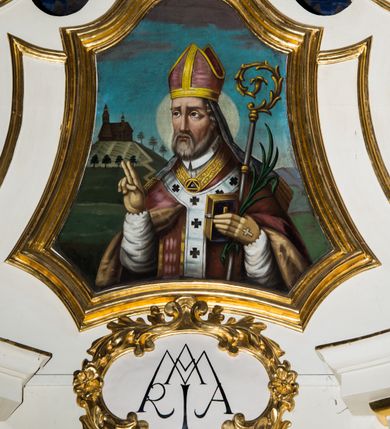 Zdjęcie nr 3: Jednokondygnacyjna i jednoosiowa nastawa ołtarza głównego uzyskała formę architektoniczną ze zwieńczeniem, ustawioną na wysokim cokole, flankowaną przez dwie bramki. Drewniany stipes ołtarzowy uzyskał kształt prostopadłościanu podtrzymującego drewnianą mensę z portatylem. Nastawa została ujęta parą opilastrowanych filarów, ustawionych pod kątem, zwróconych na zewnątrz, dźwigających pełne belkowanie przerwane nad polem środkowym oraz fragmenty przerwanego odwróconego przyczółka. Trzony pilastrów dekorowane zostały rocaille’owymi zwisami. W polu głównym znajduje się prześwit w formie stojącego prostokąta zamkniętego łukiem półkolistym nadwieszonym z umieszczonym wtórnie mniejszym obrazem Matki Boskiej z Dzieciątkiem. Nad nim usytuowano owalny kartusz ujęty ornamentem roślinnym z monogramem Marii w polu. Kondygnację flankują uszaki utworzone z ażurowych elementów rocaille. Zwieńczenie uzyskało formę trapezu o wklęsłych bokach zakończonego trzema wypustami zamkniętymi fragmentami gzymsu przechodzącego w woluty. W polu umieszczono obraz św. Wojciecha w profilowanej ramie w formie zbliżonej do trapezu o wklęsłych bokach i ściętych dolnych narożach, zamkniętego łukiem falistym. Zwieńczenie ujęte zostało liśćmi palmowymi, a zakończone muszlą i anthemionem. Bramki zostały zamknięte profilowanym gzymsem wyłamującym się po środku trójbocznie ze stojącymi na nim wazami płomienistymi. Przejścia utworzono w formie stojącego prostokąta zamkniętego łukiem półkolistym nadwieszonym. Obwiedziono je profilowaną ramą, zakończoną rozetami. Struktura nastawy została polichromowana w kolorze kości słoniowej, ze złoconymi profilami i ornamentami.