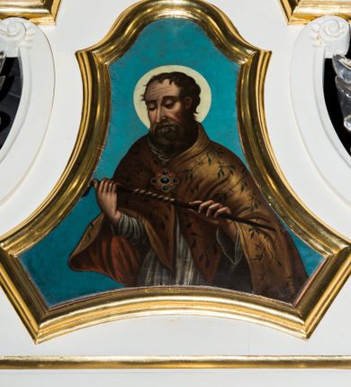 Zdjęcie nr 1: Obraz ujęty został w profilowanej ramie w formie zbliżonej do trapezu o wklęsłych bokach i ściętych dolnych narożach, zamkniętego łukiem falistym. W centrum kompozycji na niebieskim tle w półpostaci ukazano frontalnie św. Erazma. Święty przedstawiony został frontalnie z głową ujętą w trzech czwartych w lewo. Jest to starszy mężczyzna, który przed sobą w obu rękach ugiętych w łokciach trzyma metalowy pręt z nawiniętym na nim jelitem. Jego pociągła twarz o wyrazistych rysach charakteryzuje się ostrym nosem i półprzymkniętymi oczami o spojrzeniu skierowanym na trzymanym narzędziu męczeństwa. Jego głowę otaczają krótkie, zmierzwione włosy odsłaniające lewe ucho. Wokół głowy widoczny jest nimb. Erazm ubrany jest w białą komżę oraz cynobrową kapę spiętą pod szyją kosztowną fibulą dekorowaną stylizowanymi gałązkami o drobnych listkach.