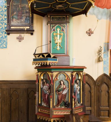 Zdjęcie nr 1: Przyścienna ambona składa się z kosza umieszczonego pierwotnie na wyższym filarze, zaplecku i baldachimu. Kosz ambony wykonano na rzucie ośmioboku, wspierając go na cokole na rzucie kwadratu przechodzącego w ośmiobok, rozszerzający się ku górze. Kosz jest artykułowany za pomocą pinakli z polami zamkniętymi łukiem ostrym z wpisanymi w nie ślepymi maswerskami. W polach znalazło się całopostaciowe przedstawienia Chrystusa oraz czterech ewangelistów. Dolna część została ujęta fryzem utworzonym z maswerków, a górna zwieńczona profilowanym parapetem. Zaplecek, opinający filar arkady tęczowej zdobi pole zamknięte łukiem ostrym z  wkomponowanym ślepym maswerkiem. W ściance zaplecka usytuowano krzyż o ramionach zakończonych trójlistnie, na tle którego znajduje się korona z krzyżującymi się palmami. W górnej części zaplecka umieszczono napis: ,,ANUNTIO VOBIS / GAUDIUM MAGNUM”. Baldachim wykonano na rzucie ośmioboku z gołębicą Ducha Świętego na podniebieniu. Wieńczy go figura Chrystusa z barankiem na ramionach. Ambona utrzymana jest w brązowo-czerwono-zielonej tonacji kolorystycznej. Dekoracja architektoniczna i ornamenty są złocone.