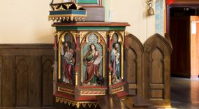 Zdjęcie nr 1: Przyścienna ambona składa się z kosza umieszczonego pierwotnie na wyższym filarze, zaplecku i baldachimu. Kosz ambony wykonano na rzucie ośmioboku, wspierając go na cokole na rzucie kwadratu przechodzącego w ośmiobok, rozszerzający się ku górze. Kosz jest artykułowany za pomocą pinakli z polami zamkniętymi łukiem ostrym z wpisanymi w nie ślepymi maswerskami. W polach znalazło się całopostaciowe przedstawienia Chrystusa oraz czterech ewangelistów. Dolna część została ujęta fryzem utworzonym z maswerków, a górna zwieńczona profilowanym parapetem. Zaplecek, opinający filar arkady tęczowej zdobi pole zamknięte łukiem ostrym z  wkomponowanym ślepym maswerkiem. W ściance zaplecka usytuowano krzyż o ramionach zakończonych trójlistnie, na tle którego znajduje się korona z krzyżującymi się palmami. W górnej części zaplecka umieszczono napis: ,,ANUNTIO VOBIS / GAUDIUM MAGNUM”. Baldachim wykonano na rzucie ośmioboku z gołębicą Ducha Świętego na podniebieniu. Wieńczy go figura Chrystusa z barankiem na ramionach. Ambona utrzymana jest w brązowo-czerwono-zielonej tonacji kolorystycznej. Dekoracja architektoniczna i ornamenty są złocone.