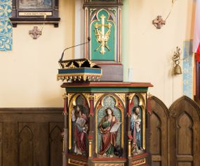 Zdjęcie nr 1: Przyścienna ambona składa się z kosza umieszczonego pierwotnie na wyższym filarze, zaplecku i baldachimu. Kosz ambony wykonano na rzucie ośmioboku, wspierając go na cokole na rzucie kwadratu przechodzącego w ośmiobok, rozszerzający się ku górze. Kosz jest artykułowany za pomocą pinakli z polami zamkniętymi łukiem ostrym z wpisanymi w nie ślepymi maswerskami. W polach znalazło się całopostaciowe przedstawienia Chrystusa oraz czterech ewangelistów. Dolna część została ujęta fryzem utworzonym z maswerków, a górna zwieńczona profilowanym parapetem. Zaplecek, opinający filar arkady tęczowej zdobi pole zamknięte łukiem ostrym z  wkomponowanym ślepym maswerkiem. W ściance zaplecka usytuowano krzyż o ramionach zakończonych trójlistnie, na tle którego znajduje się korona z krzyżującymi się palmami. W górnej części zaplecka umieszczono napis: ,,ANUNTIO VOBIS / GAUDIUM MAGNUM”. Baldachim wykonano na rzucie ośmioboku z gołębicą Ducha Świętego na podniebieniu. Wieńczy go figura Chrystusa z barankiem na ramionach. Ambona utrzymana jest w brązowo-czerwono-zielonej tonacji kolorystycznej. Dekoracja architektoniczna i ornamenty są złocone.