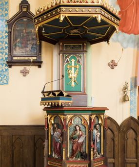 Zdjęcie nr 1: Przyścienna ambona składa się z kosza umieszczonego pierwotnie na wyższym filarze, zaplecku i baldachimu. Kosz ambony wykonano na rzucie ośmioboku, wspierając go na cokole na rzucie kwadratu przechodzącego w ośmiobok, rozszerzający się ku górze. Kosz jest artykułowany za pomocą pinakli z polami zamkniętymi łukiem ostrym z wpisanymi w nie ślepymi maswerskami. W polach znalazło się całopostaciowe przedstawienia Chrystusa oraz czterech ewangelistów. Dolna część została ujęta fryzem utworzonym z maswerków, a górna zwieńczona profilowanym parapetem. Zaplecek, opinający filar arkady tęczowej zdobi pole zamknięte łukiem ostrym z  wkomponowanym ślepym maswerkiem. W ściance zaplecka usytuowano krzyż o ramionach zakończonych trójlistnie, na tle którego znajduje się korona z krzyżującymi się palmami. W górnej części zaplecka umieszczono napis: ,,ANUNTIO VOBIS / GAUDIUM MAGNUM”. Baldachim wykonano na rzucie ośmioboku z gołębicą Ducha Świętego na podniebieniu. Wieńczy go figura Chrystusa z barankiem na ramionach. Ambona utrzymana jest w brązowo-czerwono-zielonej tonacji kolorystycznej. Dekoracja architektoniczna i ornamenty są złocone.