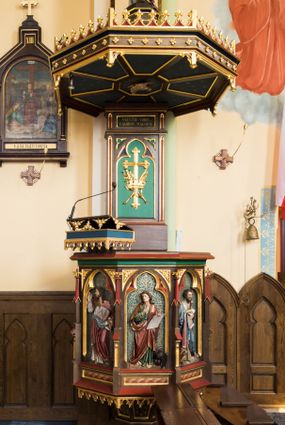 Zdjęcie nr 1: Przyścienna ambona składa się z kosza umieszczonego pierwotnie na wyższym filarze, zaplecku i baldachimu. Kosz ambony wykonano na rzucie ośmioboku, wspierając go na cokole na rzucie kwadratu przechodzącego w ośmiobok, rozszerzający się ku górze. Kosz jest artykułowany za pomocą pinakli z polami zamkniętymi łukiem ostrym z wpisanymi w nie ślepymi maswerskami. W polach znalazło się całopostaciowe przedstawienia Chrystusa oraz czterech ewangelistów. Dolna część została ujęta fryzem utworzonym z maswerków, a górna zwieńczona profilowanym parapetem. Zaplecek, opinający filar arkady tęczowej zdobi pole zamknięte łukiem ostrym z  wkomponowanym ślepym maswerkiem. W ściance zaplecka usytuowano krzyż o ramionach zakończonych trójlistnie, na tle którego znajduje się korona z krzyżującymi się palmami. W górnej części zaplecka umieszczono napis: ,,ANUNTIO VOBIS / GAUDIUM MAGNUM”. Baldachim wykonano na rzucie ośmioboku z gołębicą Ducha Świętego na podniebieniu. Wieńczy go figura Chrystusa z barankiem na ramionach. Ambona utrzymana jest w brązowo-czerwono-zielonej tonacji kolorystycznej. Dekoracja architektoniczna i ornamenty są złocone.