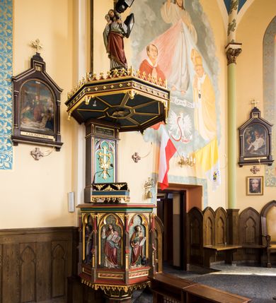 Zdjęcie nr 2: Przyścienna ambona składa się z kosza umieszczonego pierwotnie na wyższym filarze, zaplecku i baldachimu. Kosz ambony wykonano na rzucie ośmioboku, wspierając go na cokole na rzucie kwadratu przechodzącego w ośmiobok, rozszerzający się ku górze. Kosz jest artykułowany za pomocą pinakli z polami zamkniętymi łukiem ostrym z wpisanymi w nie ślepymi maswerskami. W polach znalazło się całopostaciowe przedstawienia Chrystusa oraz czterech ewangelistów. Dolna część została ujęta fryzem utworzonym z maswerków, a górna zwieńczona profilowanym parapetem. Zaplecek, opinający filar arkady tęczowej zdobi pole zamknięte łukiem ostrym z  wkomponowanym ślepym maswerkiem. W ściance zaplecka usytuowano krzyż o ramionach zakończonych trójlistnie, na tle którego znajduje się korona z krzyżującymi się palmami. W górnej części zaplecka umieszczono napis: ,,ANUNTIO VOBIS / GAUDIUM MAGNUM”. Baldachim wykonano na rzucie ośmioboku z gołębicą Ducha Świętego na podniebieniu. Wieńczy go figura Chrystusa z barankiem na ramionach. Ambona utrzymana jest w brązowo-czerwono-zielonej tonacji kolorystycznej. Dekoracja architektoniczna i ornamenty są złocone.