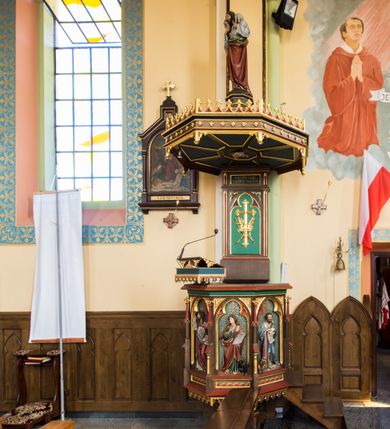 Zdjęcie nr 3: Przyścienna ambona składa się z kosza umieszczonego pierwotnie na wyższym filarze, zaplecku i baldachimu. Kosz ambony wykonano na rzucie ośmioboku, wspierając go na cokole na rzucie kwadratu przechodzącego w ośmiobok, rozszerzający się ku górze. Kosz jest artykułowany za pomocą pinakli z polami zamkniętymi łukiem ostrym z wpisanymi w nie ślepymi maswerskami. W polach znalazło się całopostaciowe przedstawienia Chrystusa oraz czterech ewangelistów. Dolna część została ujęta fryzem utworzonym z maswerków, a górna zwieńczona profilowanym parapetem. Zaplecek, opinający filar arkady tęczowej zdobi pole zamknięte łukiem ostrym z  wkomponowanym ślepym maswerkiem. W ściance zaplecka usytuowano krzyż o ramionach zakończonych trójlistnie, na tle którego znajduje się korona z krzyżującymi się palmami. W górnej części zaplecka umieszczono napis: ,,ANUNTIO VOBIS / GAUDIUM MAGNUM”. Baldachim wykonano na rzucie ośmioboku z gołębicą Ducha Świętego na podniebieniu. Wieńczy go figura Chrystusa z barankiem na ramionach. Ambona utrzymana jest w brązowo-czerwono-zielonej tonacji kolorystycznej. Dekoracja architektoniczna i ornamenty są złocone.