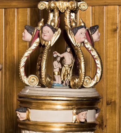 Zdjęcie nr 1: Chrzcielnica w typie kielichowym została ustawiona na prostopadłościennej, dwuczęściowej podstawie składającej się z gruszkowego trzonu oraz profilowanego cokoliku wspartym na współczesnym prostopadłościennym postumencie obłożonym drewnianą boazerią. Kolistą, spłaszczoną czarę w dolnej części ozdobiono imitacją puklowania oddzielonego za pomocą profilowanej opaski od fryzu dekorowanego uskrzydlonymi główkami aniołów i zamkniętego od góry profilowanym gzymsikiem. Pokrywę wykonano na rzucie koła wieńcząc ją ażurową strukturą utworzoną z esowato wygiętych akantowych wolut ozdobionych w górnej części główkami anielskimi. Woluty dźwigają wieniec-koronę ponad którym unosi się rzeźbiona gołębica Ducha Świętego na tle glorii promienistej i obłoków. Na samym szczycie umieszczono złocony krzyż o trójlistnie zakończonych ramionach. Wewnątrz struktury znalazła się rzeźbiona grupa przedstawiająca chrzest Chrystusa w Jordanie. Chrystusa ukazano w pozycji klęczącej z dłońmi złożonymi w geście modlitwy na piersiach. Jego biodra przepasane są złoconym perizonium. Św. Jan Chrzciciel wyobrażony został w pozycji stojącej w silnym kontrapoście. W lewej ręce trzyma laskę krzyżową, a uniesioną prawą ręką wylewa z naczynia wodę na głowę Zbawiciela. Święty ma nagi tors z luźno udrapowanym wokół ciała złotym płaszczem.
