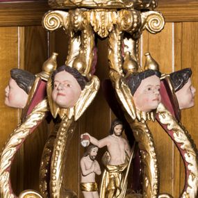 Zdjęcie nr 1: Chrzcielnica w typie kielichowym została ustawiona na prostopadłościennej, dwuczęściowej podstawie składającej się z gruszkowego trzonu oraz profilowanego cokoliku wspartym na współczesnym prostopadłościennym postumencie obłożonym drewnianą boazerią. Kolistą, spłaszczoną czarę w dolnej części ozdobiono imitacją puklowania oddzielonego za pomocą profilowanej opaski od fryzu dekorowanego uskrzydlonymi główkami aniołów i zamkniętego od góry profilowanym gzymsikiem. Pokrywę wykonano na rzucie koła wieńcząc ją ażurową strukturą utworzoną z esowato wygiętych akantowych wolut ozdobionych w górnej części główkami anielskimi. Woluty dźwigają wieniec-koronę ponad którym unosi się rzeźbiona gołębica Ducha Świętego na tle glorii promienistej i obłoków. Na samym szczycie umieszczono złocony krzyż o trójlistnie zakończonych ramionach. Wewnątrz struktury znalazła się rzeźbiona grupa przedstawiająca chrzest Chrystusa w Jordanie. Chrystusa ukazano w pozycji klęczącej z dłońmi złożonymi w geście modlitwy na piersiach. Jego biodra przepasane są złoconym perizonium. Św. Jan Chrzciciel wyobrażony został w pozycji stojącej w silnym kontrapoście. W lewej ręce trzyma laskę krzyżową, a uniesioną prawą ręką wylewa z naczynia wodę na głowę Zbawiciela. Święty ma nagi tors z luźno udrapowanym wokół ciała złotym płaszczem.
