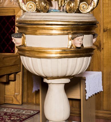 Zdjęcie nr 3: Chrzcielnica w typie kielichowym została ustawiona na prostopadłościennej, dwuczęściowej podstawie składającej się z gruszkowego trzonu oraz profilowanego cokoliku wspartym na współczesnym prostopadłościennym postumencie obłożonym drewnianą boazerią. Kolistą, spłaszczoną czarę w dolnej części ozdobiono imitacją puklowania oddzielonego za pomocą profilowanej opaski od fryzu dekorowanego uskrzydlonymi główkami aniołów i zamkniętego od góry profilowanym gzymsikiem. Pokrywę wykonano na rzucie koła wieńcząc ją ażurową strukturą utworzoną z esowato wygiętych akantowych wolut ozdobionych w górnej części główkami anielskimi. Woluty dźwigają wieniec-koronę ponad którym unosi się rzeźbiona gołębica Ducha Świętego na tle glorii promienistej i obłoków. Na samym szczycie umieszczono złocony krzyż o trójlistnie zakończonych ramionach. Wewnątrz struktury znalazła się rzeźbiona grupa przedstawiająca chrzest Chrystusa w Jordanie. Chrystusa ukazano w pozycji klęczącej z dłońmi złożonymi w geście modlitwy na piersiach. Jego biodra przepasane są złoconym perizonium. Św. Jan Chrzciciel wyobrażony został w pozycji stojącej w silnym kontrapoście. W lewej ręce trzyma laskę krzyżową, a uniesioną prawą ręką wylewa z naczynia wodę na głowę Zbawiciela. Święty ma nagi tors z luźno udrapowanym wokół ciała złotym płaszczem.
