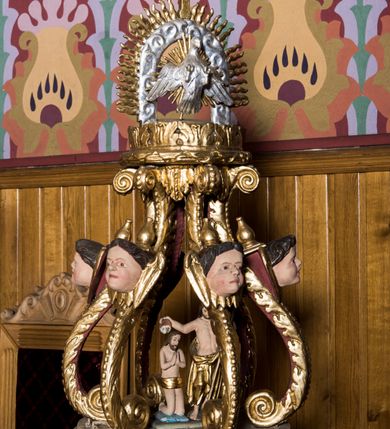 Zdjęcie nr 2: Chrzcielnica w typie kielichowym została ustawiona na prostopadłościennej, dwuczęściowej podstawie składającej się z gruszkowego trzonu oraz profilowanego cokoliku wspartym na współczesnym prostopadłościennym postumencie obłożonym drewnianą boazerią. Kolistą, spłaszczoną czarę w dolnej części ozdobiono imitacją puklowania oddzielonego za pomocą profilowanej opaski od fryzu dekorowanego uskrzydlonymi główkami aniołów i zamkniętego od góry profilowanym gzymsikiem. Pokrywę wykonano na rzucie koła wieńcząc ją ażurową strukturą utworzoną z esowato wygiętych akantowych wolut ozdobionych w górnej części główkami anielskimi. Woluty dźwigają wieniec-koronę ponad którym unosi się rzeźbiona gołębica Ducha Świętego na tle glorii promienistej i obłoków. Na samym szczycie umieszczono złocony krzyż o trójlistnie zakończonych ramionach. Wewnątrz struktury znalazła się rzeźbiona grupa przedstawiająca chrzest Chrystusa w Jordanie. Chrystusa ukazano w pozycji klęczącej z dłońmi złożonymi w geście modlitwy na piersiach. Jego biodra przepasane są złoconym perizonium. Św. Jan Chrzciciel wyobrażony został w pozycji stojącej w silnym kontrapoście. W lewej ręce trzyma laskę krzyżową, a uniesioną prawą ręką wylewa z naczynia wodę na głowę Zbawiciela. Święty ma nagi tors z luźno udrapowanym wokół ciała złotym płaszczem.
