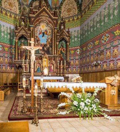 Zdjęcie nr 4: Ołtarz główny uzyskał formę architektonicznej struktury przyściennej, trójosiowej i jednokondygnacyjnej ze zwieńczeniem. Stipes ołtarzowy przyjął formę stołu, gdzie mensę z portatylem wsparto na czterech kolumnach ustawionych na wysokich wielobocznych bazach. Pomiędzy nimi usytuowano płaskorzeźbioną sceną Ostatniej Wieczerzy w kształcie leżącego prostokąta. Na mensie w osi ołtarza ustawione zostało tabernakulum z tronem wystawienia w kształcie baldachimu, stanowiące cześć środkową architektonicznego, trójosiowego założenia, gdzie w osiach bocznych w półokrągłych aediculach z wimpergami ustawiono parę adorujących aniołów. Nastawa ustawiona na wysokim cokole została dekorowana profilowanymi płycinami, ponad którymi poprowadzono profilowanym gzyms. Oś środkowa retabulum została ujęta złoconymi kolumnami o korynckich kapitelach i gładkich trzonach. Kolumny dźwigają półkoliście zamkniętą arkadę zwieńczoną profilowanym gzymsem z trójkątnie zakończoną wimpergą ozdobioną żabkami. Zwieńczenie utworzono w formie ażurowej kapliczki na rzucie wieloboku mieszczącej pełnoplastyczną figurę Michała Archanioła trzymającego miecz i wagę. Kapliczkę zakończono wieloboczną fialą o ściankach zdobionych ślepymi maswerkami. W osiach skrajnych wykonano analogiczne arkady wsparte na złoconych kolumnach mieszczące figury św. Piotra (z lewej) i św. Pawła (z prawej). W zwieńczeniach w osi znalazły się w ażurowych kapliczkach zakończonych fialami pełnoplastyczne figury św. Wojciecha i św. Stanisława w strojach pontyfikalnych. W polu ołtarza usytuowano obraz przedstawiający wszystkich świętych adorujących Trójcę Przenajświętszą. Pole ujmuje płaskorzeźbiona, złocona rama z dekoracją akantową zamknięta łukiem trójlistnym. W podłuczu wykonano arkaturę. Na zasuwie umieszczono obraz Najświętszego Serca Pana Jezusa. W płycinach wypełniających elementy architektoniczne znalazła się stylizowana dekoracja akantowa oraz motyw czwórliścia wpisanego w okrąg. Struktura ołtarza została polichromowana w odcieniach zgaszonej czerwieni i złamanego brązu wchodzącego w fiolet. Ornamentyka oraz detale elementów i profilów architektonicznych wyzłocono.