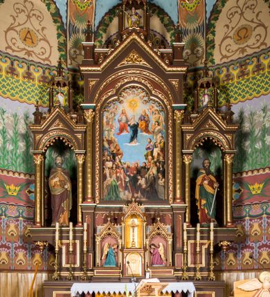 Zdjęcie nr 1: Ołtarz główny uzyskał formę architektonicznej struktury przyściennej, trójosiowej i jednokondygnacyjnej ze zwieńczeniem. Stipes ołtarzowy przyjął formę stołu, gdzie mensę z portatylem wsparto na czterech kolumnach ustawionych na wysokich wielobocznych bazach. Pomiędzy nimi usytuowano płaskorzeźbioną sceną Ostatniej Wieczerzy w kształcie leżącego prostokąta. Na mensie w osi ołtarza ustawione zostało tabernakulum z tronem wystawienia w kształcie baldachimu, stanowiące cześć środkową architektonicznego, trójosiowego założenia, gdzie w osiach bocznych w półokrągłych aediculach z wimpergami ustawiono parę adorujących aniołów. Nastawa ustawiona na wysokim cokole została dekorowana profilowanymi płycinami, ponad którymi poprowadzono profilowanym gzyms. Oś środkowa retabulum została ujęta złoconymi kolumnami o korynckich kapitelach i gładkich trzonach. Kolumny dźwigają półkoliście zamkniętą arkadę zwieńczoną profilowanym gzymsem z trójkątnie zakończoną wimpergą ozdobioną żabkami. Zwieńczenie utworzono w formie ażurowej kapliczki na rzucie wieloboku mieszczącej pełnoplastyczną figurę Michała Archanioła trzymającego miecz i wagę. Kapliczkę zakończono wieloboczną fialą o ściankach zdobionych ślepymi maswerkami. W osiach skrajnych wykonano analogiczne arkady wsparte na złoconych kolumnach mieszczące figury św. Piotra (z lewej) i św. Pawła (z prawej). W zwieńczeniach w osi znalazły się w ażurowych kapliczkach zakończonych fialami pełnoplastyczne figury św. Wojciecha i św. Stanisława w strojach pontyfikalnych. W polu ołtarza usytuowano obraz przedstawiający wszystkich świętych adorujących Trójcę Przenajświętszą. Pole ujmuje płaskorzeźbiona, złocona rama z dekoracją akantową zamknięta łukiem trójlistnym. W podłuczu wykonano arkaturę. Na zasuwie umieszczono obraz Najświętszego Serca Pana Jezusa. W płycinach wypełniających elementy architektoniczne znalazła się stylizowana dekoracja akantowa oraz motyw czwórliścia wpisanego w okrąg. Struktura ołtarza została polichromowana w odcieniach zgaszonej czerwieni i złamanego brązu wchodzącego w fiolet. Ornamentyka oraz detale elementów i profilów architektonicznych wyzłocono.