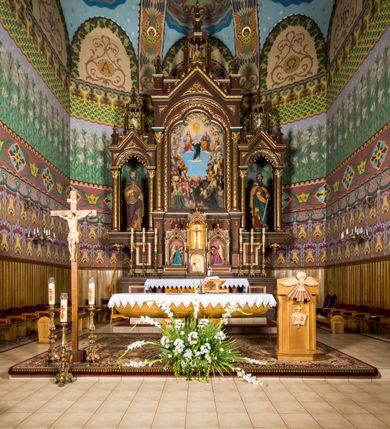 Zdjęcie nr 3: Ołtarz główny uzyskał formę architektonicznej struktury przyściennej, trójosiowej i jednokondygnacyjnej ze zwieńczeniem. Stipes ołtarzowy przyjął formę stołu, gdzie mensę z portatylem wsparto na czterech kolumnach ustawionych na wysokich wielobocznych bazach. Pomiędzy nimi usytuowano płaskorzeźbioną sceną Ostatniej Wieczerzy w kształcie leżącego prostokąta. Na mensie w osi ołtarza ustawione zostało tabernakulum z tronem wystawienia w kształcie baldachimu, stanowiące cześć środkową architektonicznego, trójosiowego założenia, gdzie w osiach bocznych w półokrągłych aediculach z wimpergami ustawiono parę adorujących aniołów. Nastawa ustawiona na wysokim cokole została dekorowana profilowanymi płycinami, ponad którymi poprowadzono profilowanym gzyms. Oś środkowa retabulum została ujęta złoconymi kolumnami o korynckich kapitelach i gładkich trzonach. Kolumny dźwigają półkoliście zamkniętą arkadę zwieńczoną profilowanym gzymsem z trójkątnie zakończoną wimpergą ozdobioną żabkami. Zwieńczenie utworzono w formie ażurowej kapliczki na rzucie wieloboku mieszczącej pełnoplastyczną figurę Michała Archanioła trzymającego miecz i wagę. Kapliczkę zakończono wieloboczną fialą o ściankach zdobionych ślepymi maswerkami. W osiach skrajnych wykonano analogiczne arkady wsparte na złoconych kolumnach mieszczące figury św. Piotra (z lewej) i św. Pawła (z prawej). W zwieńczeniach w osi znalazły się w ażurowych kapliczkach zakończonych fialami pełnoplastyczne figury św. Wojciecha i św. Stanisława w strojach pontyfikalnych. W polu ołtarza usytuowano obraz przedstawiający wszystkich świętych adorujących Trójcę Przenajświętszą. Pole ujmuje płaskorzeźbiona, złocona rama z dekoracją akantową zamknięta łukiem trójlistnym. W podłuczu wykonano arkaturę. Na zasuwie umieszczono obraz Najświętszego Serca Pana Jezusa. W płycinach wypełniających elementy architektoniczne znalazła się stylizowana dekoracja akantowa oraz motyw czwórliścia wpisanego w okrąg. Struktura ołtarza została polichromowana w odcieniach zgaszonej czerwieni i złamanego brązu wchodzącego w fiolet. Ornamentyka oraz detale elementów i profilów architektonicznych wyzłocono.