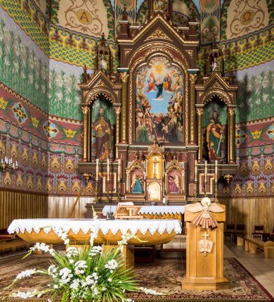 Zdjęcie nr 2: Ołtarz główny uzyskał formę architektonicznej struktury przyściennej, trójosiowej i jednokondygnacyjnej ze zwieńczeniem. Stipes ołtarzowy przyjął formę stołu, gdzie mensę z portatylem wsparto na czterech kolumnach ustawionych na wysokich wielobocznych bazach. Pomiędzy nimi usytuowano płaskorzeźbioną sceną Ostatniej Wieczerzy w kształcie leżącego prostokąta. Na mensie w osi ołtarza ustawione zostało tabernakulum z tronem wystawienia w kształcie baldachimu, stanowiące cześć środkową architektonicznego, trójosiowego założenia, gdzie w osiach bocznych w półokrągłych aediculach z wimpergami ustawiono parę adorujących aniołów. Nastawa ustawiona na wysokim cokole została dekorowana profilowanymi płycinami, ponad którymi poprowadzono profilowanym gzyms. Oś środkowa retabulum została ujęta złoconymi kolumnami o korynckich kapitelach i gładkich trzonach. Kolumny dźwigają półkoliście zamkniętą arkadę zwieńczoną profilowanym gzymsem z trójkątnie zakończoną wimpergą ozdobioną żabkami. Zwieńczenie utworzono w formie ażurowej kapliczki na rzucie wieloboku mieszczącej pełnoplastyczną figurę Michała Archanioła trzymającego miecz i wagę. Kapliczkę zakończono wieloboczną fialą o ściankach zdobionych ślepymi maswerkami. W osiach skrajnych wykonano analogiczne arkady wsparte na złoconych kolumnach mieszczące figury św. Piotra (z lewej) i św. Pawła (z prawej). W zwieńczeniach w osi znalazły się w ażurowych kapliczkach zakończonych fialami pełnoplastyczne figury św. Wojciecha i św. Stanisława w strojach pontyfikalnych. W polu ołtarza usytuowano obraz przedstawiający wszystkich świętych adorujących Trójcę Przenajświętszą. Pole ujmuje płaskorzeźbiona, złocona rama z dekoracją akantową zamknięta łukiem trójlistnym. W podłuczu wykonano arkaturę. Na zasuwie umieszczono obraz Najświętszego Serca Pana Jezusa. W płycinach wypełniających elementy architektoniczne znalazła się stylizowana dekoracja akantowa oraz motyw czwórliścia wpisanego w okrąg. Struktura ołtarza została polichromowana w odcieniach zgaszonej czerwieni i złamanego brązu wchodzącego w fiolet. Ornamentyka oraz detale elementów i profilów architektonicznych wyzłocono.