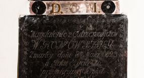 Zdjęcie nr 1: Płytowe epitafium ścienne składa się z trzech zasadniczych części - konsoli, płyty właściwej i zwieńczenia. Całość wspiera się na prostopadłościennej konsoli mającej formę leżącego, wydłużonego prostokąta. Na niej ustawiono czworoboczną płytę z inskrypcją wspartą na dwóch trapezoidalnych nóżkach, z dłuższym bokiem u góry. Płytę przy krawędziach ozdobiono ornamentem roślinnym tworzącym dekoracyjną bordiurę. Pole w całości wypełnia napis: "Magdalenie z Ostrzeszewiczów / WRONOWSKIEY / Zmarłey dnia 8[ósme]go Maia 1823 / w Roku życia 76 / przywiązane Dzieci / nayukochańszey Matce swoiey / Pomnik ten niewygasłego żalu kładą / Przechodzących / prosząc o westchnienie do Boga za Jey / Duszę". Na płycie wsparto na okrągłych podporach, pierwotnie zdobionych aplikacjami z brązu, zwieńczenie w formie płaskiego obelisku o ściętym wierzchołku. Jego pole zdobi profilowane obramienie oraz ryta arkatura. Na szczycie znajduje się półwypukła waza. Pomiędzy podporami obelisku umieszczono wykonane z brązu litery: "D[eo] O[pitmo] M[aximo]".