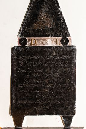 Zdjęcie nr 1: Płytowe epitafium ścienne składa się z trzech zasadniczych części - konsoli, płyty właściwej i zwieńczenia. Całość wspiera się na prostopadłościennej konsoli mającej formę leżącego, wydłużonego prostokąta. Na niej ustawiono czworoboczną płytę z inskrypcją wspartą na dwóch trapezoidalnych nóżkach, z dłuższym bokiem u góry. Płytę przy krawędziach ozdobiono ornamentem roślinnym tworzącym dekoracyjną bordiurę. Pole w całości wypełnia napis: "Magdalenie z Ostrzeszewiczów / WRONOWSKIEY / Zmarłey dnia 8[ósme]go Maia 1823 / w Roku życia 76 / przywiązane Dzieci / nayukochańszey Matce swoiey / Pomnik ten niewygasłego żalu kładą / Przechodzących / prosząc o westchnienie do Boga za Jey / Duszę". Na płycie wsparto na okrągłych podporach, pierwotnie zdobionych aplikacjami z brązu, zwieńczenie w formie płaskiego obelisku o ściętym wierzchołku. Jego pole zdobi profilowane obramienie oraz ryta arkatura. Na szczycie znajduje się półwypukła waza. Pomiędzy podporami obelisku umieszczono wykonane z brązu litery: "D[eo] O[pitmo] M[aximo]".