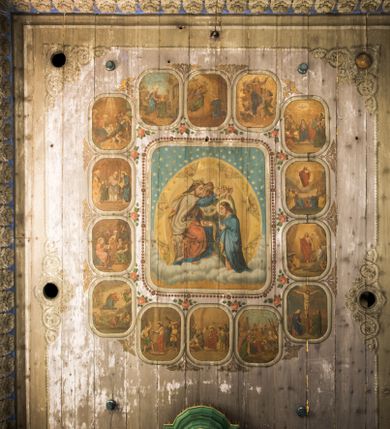 Zdjęcie nr 1: Dekoracją malarską pokryto fasety, stropy w nawie i prezbiterium, a także ich ściany oraz przestrzeń przedsionka pod wieżą. Ściany prezbiterium, nawy i przedsionka artykułowano za pomocą malowanych pilastrów o korynckich głowicach ustawionych na wysokich postumentach, podtrzymujących belkowanie. Pomiędzy pilastrami wprowadzono malowane czworoboczne płyciny o wklęsłych narożach z polem pokrytym marmoryzacją. Wąską fasetę podstropową ozdobiono fryzem z rytmicznie powtarzającym się motywem liścia palmetowego. Strop w prezbiterium i nawie uzyskał architektoniczne obramienie sceny centralnej ujętej w prostokątne pole o zaokrąglonych narożach, utworzone z iluzjonistycznie malowanych listew i zawieszonych pomiędzy nimi girland liściastych. W prezbiterium wyobrażono Trójcę Świętą, a w nawie Koronację Marii obwiedzioną tajemnicami różańcowymi w medalionach. Trójcę Świętą ukazano na obłokach na niebieskim tle. Majestatyczny Bóg Ojciec został ukazany w trzech czwartych w lewo jako siwobrody starzec o bujnych siwych włosach. W prawej ręce trzyma złote berło, lewą zaś odsuwa do siebie. Bóg Ojciec stopy wspiera o niebieski glob wyłaniający się z obłoków poniżej. Jego twarz charakteryzuje jasna karnacja, szeroko otwarte oczy, wydatny nos i usta. Głowę otacza długa, siwa broda i podobne włosy. Ubrany jest w mocno drapowaną białą tunikę i narzucony na nią rozwiany, złocisty płaszcz obszyty dekoracyjną bortą, spięty klamrą na lewym ramieniu. Obok Boga Ojca z lewej strony siedzi Chrystus, który obie, skrzyżowane nogi wspiera na globie. Zbawiciel prawą ręką podtrzymuje oparty o ramię drewniany krzyż, lewą zaś ma odsuniętą od siebie z gestem wskazującym na Ojca. Jezus został przedstawiony w trzech czwartych w prawo. Jest to dojrzały mężczyzna o pociągłej twarzy okolonej brązowymi, długimi włosami spływającymi na ramiona oraz brodą. Ma jasną karnację, prosty nos i wydatne usta. Nagi tors okrywa bladoszkarłatny płaszcz odsłaniający obie stopy Na dłoniach i stopach widoczne są rany po gwoździach. Głowę Jezusa okala świetlisty nimb. Powyżej obu postaci na tle rozjaśnionego nieba unosi się gołębica Ducha Świętego w promienistym otoku. Koronację Marii otoczoną przez owalne  medaliony ze scenami różańcowymi namalowano w polu dekoracyjnego, wydłużonego czworobocznego panneau o narożach utworzonych z roślinnego ornamentu powtórzonego symetrycznie na dłuższych bokach w środku. W centrum czworoboku o zaokrąglonych narożach obwiedzionych motywem różańca przedstawiono klęczącą na obłokach Marię przed koronującą ją Trójcą Świętą. Madonna jako młoda dziewczyna ujęta została w profilu w lewo. Lewą rękę ma przyłożoną do piersi, prawą zaś podaje synowi. Jej owalna twarz ma jasną karnację o wydatnym nosie i ustach. Głowę otaczają brązowe włosy spływające na plecy. Maria ubrana jest w lamowany złotą taśmą niebieski połaszcz oraz fragmentarycznie widoczną czerwoną suknię. Przed nią siedzi Chrystus, a za nim Bóg Ojciec. Pomiędzy nimi widać w jasnym otoku gołębicę Ducha Świętego. Jezus został ujęty w profilu w prawo. Prawą ręką unosi wraz Ojcem złotą koronę nad głową matki. Lewą zaś wspartą na kolanie trzyma jej prawą dłoń. Chrystus ma pociągłą twarz o jasnej karnacji i rumianych policzkach, wydatny nos i usta. Jego głowę otaczają długie brązowe włosy spływające na plecy oraz krótki zarost. Na głowie ma złoty diadem. Ubrany jest w niebieską tunikę i czerwony, mocno wywinięty płaszcz o jasnobeżowej podszewce. Spod tuniki widać fragmenty bosych stóp wspartych na obłokach. Obok Zbawiciela fragmentarycznie widać postać Boga Ojca ujętego frontalnie. Jest to starszy mężczyzna o siwym, obfitym zaroście i włosach. Ma wydatny nos i usta, a na głowie złoty diadem w formie mitry. Postacie ujmuje złocista, migdałowa mandorla utworzona z jasnego obramienia, na którym ukazano uskrzydlone główki serafinów. Dolną część mandorli spowijają obłoki. Całość ukazano na niebieskim tle usianym gwiazdami. Wokół centralnego przedstawienia rozmieszczono czternaście owalnych medalionów różańcowych. Ich narracja rozpoczyna się na osi ponad sceną z koronacją Marii. Idąc w lewo namalowano: Zwiastowanie Marii, Nawiedzenie św. Elżbiety, Boże Narodzenie, Ofiarowanie w świątyni, Znalezienie w świątyni, Modlitwa w Ogrójcu, Biczowanie, Cierniem ukoronowanie, Niesienie krzyża, Ukrzyżowanie, Zmartwychwstanie, Wniebowstąpienie, Zesłanie Ducha Świętego i Wniebowzięcie Marii. Pomiędzy medalionami namalowano czerwone róże.    