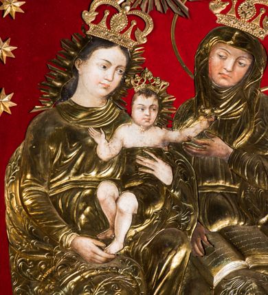 Zdjęcie nr 7: Obraz uzyskał kształt stojącego prostokąta zamkniętego półkoliście. Jego tło przykryto czerwonym aksamitem, na którym zamocowano 16 metalowych, złoconych gwiazd, rozmieszczonych wzdłuż arkady. Postacie ozdobiono trybowanymi, metalowymi i złoconymi sukienkami wotywnymi z koronami, odsłaniającymi partie twarzy, rąk i stóp Marii i św. Anny oraz ciała Dzieciątka. W centrum kompozycji na ławie, obok siebie siedzi Matka Boska z Dzieciątkiem i św. Anna. Siedząca z lewej strony Madonna ukazana została w całej postaci w ujęciu trzech czwartych w prawo. Maria lewą ręką podtrzymuje Dzieciątko za tors, a prawą za lewą stopę. Madonna ma owalną twarz o pucołowatych, rumianych policzkach z prostym nosem, otwartymi oczami i wydatnymi ustami. Wokół głowy widoczne są brązowe włosy, zaczesane za prawe ucho i spływające na ramiona. Mały Jezus ma rozłożone ręce. Jego postać została ujęta frontalnie z głową w trzech czwarty w prawo. Prawą rękę ma zgiętą w łokciu i skierowaną w stronę Matki, a lewą sięga w stronę św. Anny. Zbawiciel ma owalną twarz o pucułowatych, rumianych policzkach z drobnym nosem i ustami. Jego biodra okrywa tkanina przykryta sukienką wotywną. Głowę Marii i Dzieciątka otaczają metalowe, złocone, promieniste niby. Święta Anna została przedstawiona w całej postaci w ujęciu trzech czwartych w prawo. Prawą ręką trzyma na kolanach rozłożoną księgę, a lewą unosi w stronę Dzieciątka. Ponad głowami Marii i Anny przedstawiono gołębicę Ducha Świętego z rozpostartymi skrzydłami, otoczoną metalowymi i złoconymi promieniami.