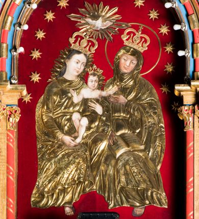 Zdjęcie nr 1: Obraz uzyskał kształt stojącego prostokąta zamkniętego półkoliście. Jego tło przykryto czerwonym aksamitem, na którym zamocowano 16 metalowych, złoconych gwiazd, rozmieszczonych wzdłuż arkady. Postacie ozdobiono trybowanymi, metalowymi i złoconymi sukienkami wotywnymi z koronami, odsłaniającymi partie twarzy, rąk i stóp Marii i św. Anny oraz ciała Dzieciątka. W centrum kompozycji na ławie, obok siebie siedzi Matka Boska z Dzieciątkiem i św. Anna. Siedząca z lewej strony Madonna ukazana została w całej postaci w ujęciu trzech czwartych w prawo. Maria lewą ręką podtrzymuje Dzieciątko za tors, a prawą za lewą stopę. Madonna ma owalną twarz o pucołowatych, rumianych policzkach z prostym nosem, otwartymi oczami i wydatnymi ustami. Wokół głowy widoczne są brązowe włosy, zaczesane za prawe ucho i spływające na ramiona. Mały Jezus ma rozłożone ręce. Jego postać została ujęta frontalnie z głową w trzech czwarty w prawo. Prawą rękę ma zgiętą w łokciu i skierowaną w stronę Matki, a lewą sięga w stronę św. Anny. Zbawiciel ma owalną twarz o pucułowatych, rumianych policzkach z drobnym nosem i ustami. Jego biodra okrywa tkanina przykryta sukienką wotywną. Głowę Marii i Dzieciątka otaczają metalowe, złocone, promieniste niby. Święta Anna została przedstawiona w całej postaci w ujęciu trzech czwartych w prawo. Prawą ręką trzyma na kolanach rozłożoną księgę, a lewą unosi w stronę Dzieciątka. Ponad głowami Marii i Anny przedstawiono gołębicę Ducha Świętego z rozpostartymi skrzydłami, otoczoną metalowymi i złoconymi promieniami.