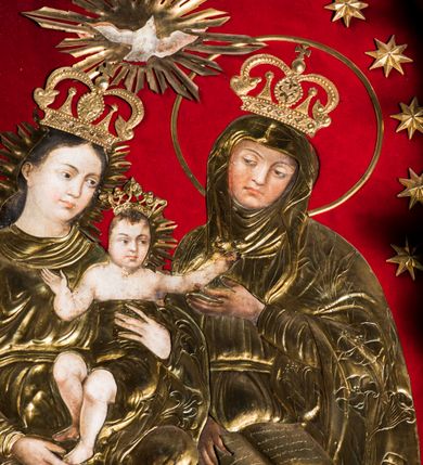 Zdjęcie nr 3: Obraz uzyskał kształt stojącego prostokąta zamkniętego półkoliście. Jego tło przykryto czerwonym aksamitem, na którym zamocowano 16 metalowych, złoconych gwiazd, rozmieszczonych wzdłuż arkady. Postacie ozdobiono trybowanymi, metalowymi i złoconymi sukienkami wotywnymi z koronami, odsłaniającymi partie twarzy, rąk i stóp Marii i św. Anny oraz ciała Dzieciątka. W centrum kompozycji na ławie, obok siebie siedzi Matka Boska z Dzieciątkiem i św. Anna. Siedząca z lewej strony Madonna ukazana została w całej postaci w ujęciu trzech czwartych w prawo. Maria lewą ręką podtrzymuje Dzieciątko za tors, a prawą za lewą stopę. Madonna ma owalną twarz o pucołowatych, rumianych policzkach z prostym nosem, otwartymi oczami i wydatnymi ustami. Wokół głowy widoczne są brązowe włosy, zaczesane za prawe ucho i spływające na ramiona. Mały Jezus ma rozłożone ręce. Jego postać została ujęta frontalnie z głową w trzech czwarty w prawo. Prawą rękę ma zgiętą w łokciu i skierowaną w stronę Matki, a lewą sięga w stronę św. Anny. Zbawiciel ma owalną twarz o pucułowatych, rumianych policzkach z drobnym nosem i ustami. Jego biodra okrywa tkanina przykryta sukienką wotywną. Głowę Marii i Dzieciątka otaczają metalowe, złocone, promieniste niby. Święta Anna została przedstawiona w całej postaci w ujęciu trzech czwartych w prawo. Prawą ręką trzyma na kolanach rozłożoną księgę, a lewą unosi w stronę Dzieciątka. Ponad głowami Marii i Anny przedstawiono gołębicę Ducha Świętego z rozpostartymi skrzydłami, otoczoną metalowymi i złoconymi promieniami.