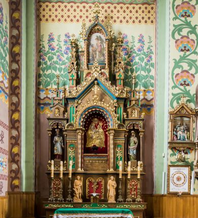 Zdjęcie nr 1: Trójosiowy ołtarz przyścienny uzyskał formę architektonicznej nastawy jednokondygnacyjnej ze zwieńczeniem. Sarkofagowy stipes ołtarzowy wsparto na profilowanym cokole, wieńcząc go gzymsem. Wspiera on drewnianą mensę z portatylem. Ołtarz ustawiono na podeście z dwoma gradusami. Retabulum zostało zaopatrzone w wysoki, drewniany cokół zwieńczony dekoracyjną listwą z motywami roślinno-kwiatowymi. Na cokole ustawiono trójosiową predellę. Jej środkową część wydzielono za pomocą wysokich, fazowanych postumentów, pomiędzy którymi znalazła się płycina z połączonymi ze sobą dwiema arkadami, których łuki przechodzą na profilowany wspornik ozdobiony ornamentem roślinno-kwiatowym. Płaskorzeźbiony ornament roślinno-kwiatowy poprowadzono także w świetle arkady. Po bokach widoczne są dwie kolejne, półkoliste arkady z podłuczami wypełnionymi arkaturą o czerwonych polach z płaskorzeźbionym ornamentem kwiatowo-roślinnym. W kondygnacji na osi środkowej usytuowano półkolistą arkadę ze zdwojonymi archiwoltami, wspartymi na podwójnych półkolumnach, o prostych bazach i kapitelach z motywem roślinnym. Arkadę flankują postumenty z półkoliście zamkniętymi płycinami o zielonych polach, wypełnionych płaskorzeźbionymi, skrzydlatymi główkami aniołków i motywami roślinnymi. Ponad gzymsem znalazły się półkoliste muszle. Arkadę zdobi biegnący po zewnętrznej ornament utworzony ze stylizowanych liści oraz szyszek, przedzielony na osi krzyżem. We wnętrz arkady umieszczono obraz w kształcie prostokąta zakończony półkoliście z przedstawieniem św. Anny Samotrzeć, dekorowany sukienką wotywną, nałożoną na aksamitne tło. Obraz zasłaniają dwie zasuwy z przedstawieniem Matki Boskiej Nieustającej Pomocy i św. Józefa. Poniżej obrazu widoczny jest parapet cokołu wprowadzonego w dolnej części arkady. Arkadę wieńczy belkowanie wyłamane na osi w formie nadwieszonego trójkąta, wsparte na dwóch  postumentach. Szczyt dekoruje ornament roślinny z motywem kół oraz ćwierci półkolistych muszli i stylizowany neogotycki kwiaton. Postumenty, wypełnione zaślepionym maswerkiem, ponad gzymsem przechodzą w dwie czworoboczne fiale, zakończone ostrosłupowym zwieńczeniem dekorowanym żabkami i kwiatonami, przechodzącymi w krzyże o trójlistnie zakończonych ramionach. W dolnych częściach fial usytuowano wycinane, trójlistne, zielone płyciny z motywem koniczyny przechodzące w listewki zakończone trójlistnie. Pomiędzy fialami wypiętrzono zwieńczenie utworzone z ostrołukowej arkady wspartej na zdwojonych półkolumnach. Na szczycie arkady umieszczono stylizowany neogotycki kwiaton, a po bokach płaskorzeźbione ornamenty kwiatowo-roślinne. Pod arkadą w polu zwieńczenia umieszczono prostokątny obraz zamknięty półkoliście z przedstawieniem Archanioła Rafała i Tobiasza. W osiach bocznych retabulum na poziomie kondygnacji znalazły się dwie wnęki arkadowe wsparte na kolumnach o roślinnych kapitelach i prostych bazach, umieszczonych na niskich cokołach. Całość została wsparta na cokoliku dekorowanym ornamentem roślinnym. Półkoliste arkady wypełniono pseudogotyckim maswerkiem. We wnękach ustawiono pełnoplastyczne rzeźby św. Zachariasza i św. Elżbiety. Powyżej usytuowano dekoracyjne zwieńczenie nawiązujące do pseudogotyckich fial z trzema sterczynami.