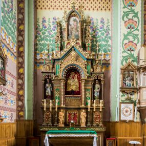Zdjęcie nr 1: Trójosiowy ołtarz przyścienny uzyskał formę architektonicznej nastawy jednokondygnacyjnej ze zwieńczeniem. Sarkofagowy stipes ołtarzowy wsparto na profilowanym cokole, wieńcząc go gzymsem. Wspiera on drewnianą mensę z portatylem. Ołtarz ustawiono na podeście z dwoma gradusami. Retabulum zostało zaopatrzone w wysoki, drewniany cokół zwieńczony dekoracyjną listwą z motywami roślinno-kwiatowymi. Na cokole ustawiono trójosiową predellę. Jej środkową część wydzielono za pomocą wysokich, fazowanych postumentów, pomiędzy którymi znalazła się płycina z połączonymi ze sobą dwiema arkadami, których łuki przechodzą na profilowany wspornik ozdobiony ornamentem roślinno-kwiatowym. Płaskorzeźbiony ornament roślinno-kwiatowy poprowadzono także w świetle arkady. Po bokach widoczne są dwie kolejne, półkoliste arkady z podłuczami wypełnionymi arkaturą o czerwonych polach z płaskorzeźbionym ornamentem kwiatowo-roślinnym. W kondygnacji na osi środkowej usytuowano półkolistą arkadę ze zdwojonymi archiwoltami, wspartymi na podwójnych półkolumnach, o prostych bazach i kapitelach z motywem roślinnym. Arkadę flankują postumenty z półkoliście zamkniętymi płycinami o zielonych polach, wypełnionych płaskorzeźbionymi, skrzydlatymi główkami aniołków i motywami roślinnymi. Ponad gzymsem znalazły się półkoliste muszle. Arkadę zdobi biegnący po zewnętrznej ornament utworzony ze stylizowanych liści oraz szyszek, przedzielony na osi krzyżem. We wnętrz arkady umieszczono obraz w kształcie prostokąta zakończony półkoliście z przedstawieniem św. Anny Samotrzeć, dekorowany sukienką wotywną, nałożoną na aksamitne tło. Obraz zasłaniają dwie zasuwy z przedstawieniem Matki Boskiej Nieustającej Pomocy i św. Józefa. Poniżej obrazu widoczny jest parapet cokołu wprowadzonego w dolnej części arkady. Arkadę wieńczy belkowanie wyłamane na osi w formie nadwieszonego trójkąta, wsparte na dwóch  postumentach. Szczyt dekoruje ornament roślinny z motywem kół oraz ćwierci półkolistych muszli i stylizowany neogotycki kwiaton. Postumenty, wypełnione zaślepionym maswerkiem, ponad gzymsem przechodzą w dwie czworoboczne fiale, zakończone ostrosłupowym zwieńczeniem dekorowanym żabkami i kwiatonami, przechodzącymi w krzyże o trójlistnie zakończonych ramionach. W dolnych częściach fial usytuowano wycinane, trójlistne, zielone płyciny z motywem koniczyny przechodzące w listewki zakończone trójlistnie. Pomiędzy fialami wypiętrzono zwieńczenie utworzone z ostrołukowej arkady wspartej na zdwojonych półkolumnach. Na szczycie arkady umieszczono stylizowany neogotycki kwiaton, a po bokach płaskorzeźbione ornamenty kwiatowo-roślinne. Pod arkadą w polu zwieńczenia umieszczono prostokątny obraz zamknięty półkoliście z przedstawieniem Archanioła Rafała i Tobiasza. W osiach bocznych retabulum na poziomie kondygnacji znalazły się dwie wnęki arkadowe wsparte na kolumnach o roślinnych kapitelach i prostych bazach, umieszczonych na niskich cokołach. Całość została wsparta na cokoliku dekorowanym ornamentem roślinnym. Półkoliste arkady wypełniono pseudogotyckim maswerkiem. We wnękach ustawiono pełnoplastyczne rzeźby św. Zachariasza i św. Elżbiety. Powyżej usytuowano dekoracyjne zwieńczenie nawiązujące do pseudogotyckich fial z trzema sterczynami.