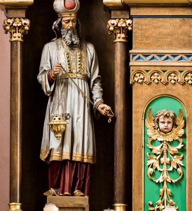 Zdjęcie nr 10: Trójosiowy ołtarz przyścienny uzyskał formę architektonicznej nastawy jednokondygnacyjnej ze zwieńczeniem. Sarkofagowy stipes ołtarzowy wsparto na profilowanym cokole, wieńcząc go gzymsem. Wspiera on drewnianą mensę z portatylem. Ołtarz ustawiono na podeście z dwoma gradusami. Retabulum zostało zaopatrzone w wysoki, drewniany cokół zwieńczony dekoracyjną listwą z motywami roślinno-kwiatowymi. Na cokole ustawiono trójosiową predellę. Jej środkową część wydzielono za pomocą wysokich, fazowanych postumentów, pomiędzy którymi znalazła się płycina z połączonymi ze sobą dwiema arkadami, których łuki przechodzą na profilowany wspornik ozdobiony ornamentem roślinno-kwiatowym. Płaskorzeźbiony ornament roślinno-kwiatowy poprowadzono także w świetle arkady. Po bokach widoczne są dwie kolejne, półkoliste arkady z podłuczami wypełnionymi arkaturą o czerwonych polach z płaskorzeźbionym ornamentem kwiatowo-roślinnym. W kondygnacji na osi środkowej usytuowano półkolistą arkadę ze zdwojonymi archiwoltami, wspartymi na podwójnych półkolumnach, o prostych bazach i kapitelach z motywem roślinnym. Arkadę flankują postumenty z półkoliście zamkniętymi płycinami o zielonych polach, wypełnionych płaskorzeźbionymi, skrzydlatymi główkami aniołków i motywami roślinnymi. Ponad gzymsem znalazły się półkoliste muszle. Arkadę zdobi biegnący po zewnętrznej ornament utworzony ze stylizowanych liści oraz szyszek, przedzielony na osi krzyżem. We wnętrz arkady umieszczono obraz w kształcie prostokąta zakończony półkoliście z przedstawieniem św. Anny Samotrzeć, dekorowany sukienką wotywną, nałożoną na aksamitne tło. Obraz zasłaniają dwie zasuwy z przedstawieniem Matki Boskiej Nieustającej Pomocy i św. Józefa. Poniżej obrazu widoczny jest parapet cokołu wprowadzonego w dolnej części arkady. Arkadę wieńczy belkowanie wyłamane na osi w formie nadwieszonego trójkąta, wsparte na dwóch  postumentach. Szczyt dekoruje ornament roślinny z motywem kół oraz ćwierci półkolistych muszli i stylizowany neogotycki kwiaton. Postumenty, wypełnione zaślepionym maswerkiem, ponad gzymsem przechodzą w dwie czworoboczne fiale, zakończone ostrosłupowym zwieńczeniem dekorowanym żabkami i kwiatonami, przechodzącymi w krzyże o trójlistnie zakończonych ramionach. W dolnych częściach fial usytuowano wycinane, trójlistne, zielone płyciny z motywem koniczyny przechodzące w listewki zakończone trójlistnie. Pomiędzy fialami wypiętrzono zwieńczenie utworzone z ostrołukowej arkady wspartej na zdwojonych półkolumnach. Na szczycie arkady umieszczono stylizowany neogotycki kwiaton, a po bokach płaskorzeźbione ornamenty kwiatowo-roślinne. Pod arkadą w polu zwieńczenia umieszczono prostokątny obraz zamknięty półkoliście z przedstawieniem Archanioła Rafała i Tobiasza. W osiach bocznych retabulum na poziomie kondygnacji znalazły się dwie wnęki arkadowe wsparte na kolumnach o roślinnych kapitelach i prostych bazach, umieszczonych na niskich cokołach. Całość została wsparta na cokoliku dekorowanym ornamentem roślinnym. Półkoliste arkady wypełniono pseudogotyckim maswerkiem. We wnękach ustawiono pełnoplastyczne rzeźby św. Zachariasza i św. Elżbiety. Powyżej usytuowano dekoracyjne zwieńczenie nawiązujące do pseudogotyckich fial z trzema sterczynami.