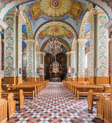 Zdjęcie nr 6: Dekoracja malarska wnętrza kościoła została oparta zasadniczo na motywach ornamentalnych, gdzie wyjątek stanowi monumentalna scena ukazująca uczniów idących do Emaus, usytuowana na północnej ścianie prezbiterium, nad wejściem do zakrystii. Wyobrażono tu w malowanym, ornamentalnym obramieniu scenę wpisaną w stojący prostokąt, zamknięty od góry łukiem półkolistym. W centrum kompozycji ukazano Chrystusa, którego zatrzymują stojący na schodach gospody dwaj uczniowie idący do Emaus. Stojący Zbawiciel został wyobrażony z lewej strony w trzech czwartych w prawo. Prawą ręką podtrzymuje połę płaszcza, lewa jest niewidoczna. Jest to dojrzały mężczyzna o pociągłej twarzy z wydatnym nosem i ustami, długimi włosami spływającymi na ramiona oraz brodą okalającą twarz o jasnej karnacji. Zbawiciel ubrany jest w białą tunikę i szkarłatny płaszcz. Do niego zwraca się jedne z uczniów ukazany tyłem do widza w trzech czwartych w lewo. Jest to dojrzały mężczyzna, który lewą rękę wyciąga w stronę Jezusa, prawą zaś zdaje się zapraszać do wejścia na schody. Jego twarz okolona krótkimi włosami z brodą i wydatnym nosem i ustami widoczna jest z profilu. Ubrany jest w jasną tunikę oraz jasnobeżowy płaszcz. Przed nim z prawej strony na stopniach wiodących do drzwi gospody stoi drugi uczeń. Ujęto go frontalnie w kontrapoście z głową w trzech czwartych w lewo. W prawej ręce trzyma laskę, lewą zaś wskazuje na drzwi zajazdu. Jest to młody mężczyzna o pociągłej twarzy i długich włosach spływających na plecy. Ubrany jest w przewiązaną w pasie tunikę oraz narzucony na ramiona bladoszkarłatny płaszcz. Za przedstawionymi postaciami widać fragment wschodniej architektury oraz rozbudowaną sekwencję pejzażową z elementem bramy miejskiej oraz roślinnością na tle wieczornego nieba. Poniżej przedstawienia umieszczono napis: "Uczniowie z Emaus" podpis: "ZOSTAŃ Z NAMI, BOĆ SIĘ MA KU WIECZOROWI I DZIEŃ SIĘ JUŻ NACHYLIŁ - ŚW[IĘTY] ŁUKASZ XXIV". Bogactwo ornamentalnej dekoracji malarskiej zostało podporządkowane architekturze wnętrza. Partię ścian pokryto malaturą o charakterze dywanowym, ułożoną strefowo z powtarzających się rytmicznie kolorowych motywów ornamentalnych. Gurty sklepienne oraz trzony pilastrów na filarach międzynawowych pokryto wykonanymi na niebieskim lub błękitnym tle ornamentalnymi pasami z motywem fantazyjnych kwiatów i wici roślinnych. Partię sklepienne w kolejnych przęsłach w nawach udekorowano koncentrycznie rozchodzą się płomienistą i falującą ornamentyką, z wplecionymi motywami symbolu Najświętszego Serca Pana Jezusa, Apokaliptycznego Baranka, kielicha z hostią w otoczeniu kłosów zbóż i winnej latorośli, a także gałązek różanych i cierniowych. Na ścianach prezbiterium zastosowano podział strefowy. Dolne przestrzenie ozdobiono pasma ornamentalnymi. Powyżej rozmieszczono strefę z przedstawieniem rosnących obok siebie kwitów lilii. U samej góry sięgnięto do motywów rybich łusek oraz stylizowanej, wielobarwnej ornamentyki fantazyjnej. Gurty sklepienia udekorowano pasami opartymi na stylizowanych skrzydłach serafinów. Natomiast w poszczególnych polach sklepiennych na niebieskim tle w fantazyjnych, falujących otokach wyobrażono dwa skrzyżowane płonące miecze, oko Opatrzności Bożej, dwie tablice Dekalogu. Na środkowej gurcie u szczytu w barwnym obramienium ukazano napis: "KTÓŻ JAK BÓG". 