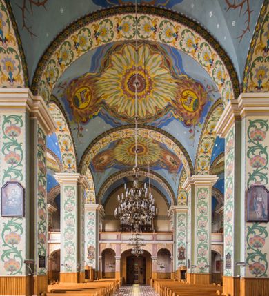 Zdjęcie nr 1: Dekoracja malarska wnętrza kościoła została oparta zasadniczo na motywach ornamentalnych, gdzie wyjątek stanowi monumentalna scena ukazująca uczniów idących do Emaus, usytuowana na północnej ścianie prezbiterium, nad wejściem do zakrystii. Wyobrażono tu w malowanym, ornamentalnym obramieniu scenę wpisaną w stojący prostokąt, zamknięty od góry łukiem półkolistym. W centrum kompozycji ukazano Chrystusa, którego zatrzymują stojący na schodach gospody dwaj uczniowie idący do Emaus. Stojący Zbawiciel został wyobrażony z lewej strony w trzech czwartych w prawo. Prawą ręką podtrzymuje połę płaszcza, lewa jest niewidoczna. Jest to dojrzały mężczyzna o pociągłej twarzy z wydatnym nosem i ustami, długimi włosami spływającymi na ramiona oraz brodą okalającą twarz o jasnej karnacji. Zbawiciel ubrany jest w białą tunikę i szkarłatny płaszcz. Do niego zwraca się jedne z uczniów ukazany tyłem do widza w trzech czwartych w lewo. Jest to dojrzały mężczyzna, który lewą rękę wyciąga w stronę Jezusa, prawą zaś zdaje się zapraszać do wejścia na schody. Jego twarz okolona krótkimi włosami z brodą i wydatnym nosem i ustami widoczna jest z profilu. Ubrany jest w jasną tunikę oraz jasnobeżowy płaszcz. Przed nim z prawej strony na stopniach wiodących do drzwi gospody stoi drugi uczeń. Ujęto go frontalnie w kontrapoście z głową w trzech czwartych w lewo. W prawej ręce trzyma laskę, lewą zaś wskazuje na drzwi zajazdu. Jest to młody mężczyzna o pociągłej twarzy i długich włosach spływających na plecy. Ubrany jest w przewiązaną w pasie tunikę oraz narzucony na ramiona bladoszkarłatny płaszcz. Za przedstawionymi postaciami widać fragment wschodniej architektury oraz rozbudowaną sekwencję pejzażową z elementem bramy miejskiej oraz roślinnością na tle wieczornego nieba. Poniżej przedstawienia umieszczono napis: &quot;Uczniowie z Emaus&quot; podpis: &quot;ZOSTAŃ Z NAMI, BOĆ SIĘ MA KU WIECZOROWI I DZIEŃ SIĘ JUŻ NACHYLIŁ - ŚW[IĘTY] ŁUKASZ XXIV&quot;. Bogactwo ornamentalnej dekoracji malarskiej zostało podporządkowane architekturze wnętrza. Partię ścian pokryto malaturą o charakterze dywanowym, ułożoną strefowo z powtarzających się rytmicznie kolorowych motywów ornamentalnych. Gurty sklepienne oraz trzony pilastrów na filarach międzynawowych pokryto wykonanymi na niebieskim lub błękitnym tle ornamentalnymi pasami z motywem fantazyjnych kwiatów i wici roślinnych. Partię sklepienne w kolejnych przęsłach w nawach udekorowano koncentrycznie rozchodzą się płomienistą i falującą ornamentyką, z wplecionymi motywami symbolu Najświętszego Serca Pana Jezusa, Apokaliptycznego Baranka, kielicha z hostią w otoczeniu kłosów zbóż i winnej latorośli, a także gałązek różanych i cierniowych. Na ścianach prezbiterium zastosowano podział strefowy. Dolne przestrzenie ozdobiono pasma ornamentalnymi. Powyżej rozmieszczono strefę z przedstawieniem rosnących obok siebie kwitów lilii. U samej góry sięgnięto do motywów rybich łusek oraz stylizowanej, wielobarwnej ornamentyki fantazyjnej. Gurty sklepienia udekorowano pasami opartymi na stylizowanych skrzydłach serafinów. Natomiast w poszczególnych polach sklepiennych na niebieskim tle w fantazyjnych, falujących otokach wyobrażono dwa skrzyżowane płonące miecze, oko Opatrzności Bożej, dwie tablice Dekalogu. Na środkowej gurcie u szczytu w barwnym obramienium ukazano napis: &quot;KTÓŻ JAK BÓG&quot;. 