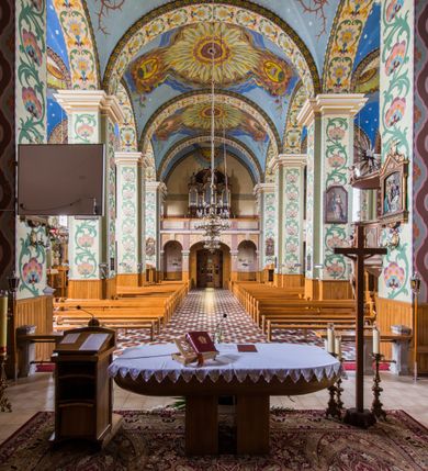 Zdjęcie nr 7: Dekoracja malarska wnętrza kościoła została oparta zasadniczo na motywach ornamentalnych, gdzie wyjątek stanowi monumentalna scena ukazująca uczniów idących do Emaus, usytuowana na północnej ścianie prezbiterium, nad wejściem do zakrystii. Wyobrażono tu w malowanym, ornamentalnym obramieniu scenę wpisaną w stojący prostokąt, zamknięty od góry łukiem półkolistym. W centrum kompozycji ukazano Chrystusa, którego zatrzymują stojący na schodach gospody dwaj uczniowie idący do Emaus. Stojący Zbawiciel został wyobrażony z lewej strony w trzech czwartych w prawo. Prawą ręką podtrzymuje połę płaszcza, lewa jest niewidoczna. Jest to dojrzały mężczyzna o pociągłej twarzy z wydatnym nosem i ustami, długimi włosami spływającymi na ramiona oraz brodą okalającą twarz o jasnej karnacji. Zbawiciel ubrany jest w białą tunikę i szkarłatny płaszcz. Do niego zwraca się jedne z uczniów ukazany tyłem do widza w trzech czwartych w lewo. Jest to dojrzały mężczyzna, który lewą rękę wyciąga w stronę Jezusa, prawą zaś zdaje się zapraszać do wejścia na schody. Jego twarz okolona krótkimi włosami z brodą i wydatnym nosem i ustami widoczna jest z profilu. Ubrany jest w jasną tunikę oraz jasnobeżowy płaszcz. Przed nim z prawej strony na stopniach wiodących do drzwi gospody stoi drugi uczeń. Ujęto go frontalnie w kontrapoście z głową w trzech czwartych w lewo. W prawej ręce trzyma laskę, lewą zaś wskazuje na drzwi zajazdu. Jest to młody mężczyzna o pociągłej twarzy i długich włosach spływających na plecy. Ubrany jest w przewiązaną w pasie tunikę oraz narzucony na ramiona bladoszkarłatny płaszcz. Za przedstawionymi postaciami widać fragment wschodniej architektury oraz rozbudowaną sekwencję pejzażową z elementem bramy miejskiej oraz roślinnością na tle wieczornego nieba. Poniżej przedstawienia umieszczono napis: "Uczniowie z Emaus" podpis: "ZOSTAŃ Z NAMI, BOĆ SIĘ MA KU WIECZOROWI I DZIEŃ SIĘ JUŻ NACHYLIŁ - ŚW[IĘTY] ŁUKASZ XXIV". Bogactwo ornamentalnej dekoracji malarskiej zostało podporządkowane architekturze wnętrza. Partię ścian pokryto malaturą o charakterze dywanowym, ułożoną strefowo z powtarzających się rytmicznie kolorowych motywów ornamentalnych. Gurty sklepienne oraz trzony pilastrów na filarach międzynawowych pokryto wykonanymi na niebieskim lub błękitnym tle ornamentalnymi pasami z motywem fantazyjnych kwiatów i wici roślinnych. Partię sklepienne w kolejnych przęsłach w nawach udekorowano koncentrycznie rozchodzą się płomienistą i falującą ornamentyką, z wplecionymi motywami symbolu Najświętszego Serca Pana Jezusa, Apokaliptycznego Baranka, kielicha z hostią w otoczeniu kłosów zbóż i winnej latorośli, a także gałązek różanych i cierniowych. Na ścianach prezbiterium zastosowano podział strefowy. Dolne przestrzenie ozdobiono pasma ornamentalnymi. Powyżej rozmieszczono strefę z przedstawieniem rosnących obok siebie kwitów lilii. U samej góry sięgnięto do motywów rybich łusek oraz stylizowanej, wielobarwnej ornamentyki fantazyjnej. Gurty sklepienia udekorowano pasami opartymi na stylizowanych skrzydłach serafinów. Natomiast w poszczególnych polach sklepiennych na niebieskim tle w fantazyjnych, falujących otokach wyobrażono dwa skrzyżowane płonące miecze, oko Opatrzności Bożej, dwie tablice Dekalogu. Na środkowej gurcie u szczytu w barwnym obramienium ukazano napis: "KTÓŻ JAK BÓG". 