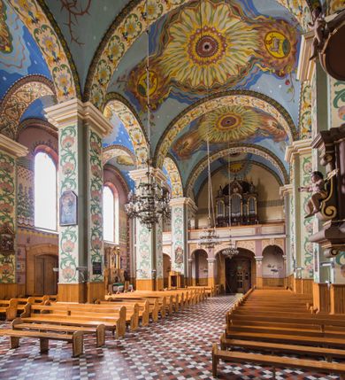 Zdjęcie nr 9: Dekoracja malarska wnętrza kościoła została oparta zasadniczo na motywach ornamentalnych, gdzie wyjątek stanowi monumentalna scena ukazująca uczniów idących do Emaus, usytuowana na północnej ścianie prezbiterium, nad wejściem do zakrystii. Wyobrażono tu w malowanym, ornamentalnym obramieniu scenę wpisaną w stojący prostokąt, zamknięty od góry łukiem półkolistym. W centrum kompozycji ukazano Chrystusa, którego zatrzymują stojący na schodach gospody dwaj uczniowie idący do Emaus. Stojący Zbawiciel został wyobrażony z lewej strony w trzech czwartych w prawo. Prawą ręką podtrzymuje połę płaszcza, lewa jest niewidoczna. Jest to dojrzały mężczyzna o pociągłej twarzy z wydatnym nosem i ustami, długimi włosami spływającymi na ramiona oraz brodą okalającą twarz o jasnej karnacji. Zbawiciel ubrany jest w białą tunikę i szkarłatny płaszcz. Do niego zwraca się jedne z uczniów ukazany tyłem do widza w trzech czwartych w lewo. Jest to dojrzały mężczyzna, który lewą rękę wyciąga w stronę Jezusa, prawą zaś zdaje się zapraszać do wejścia na schody. Jego twarz okolona krótkimi włosami z brodą i wydatnym nosem i ustami widoczna jest z profilu. Ubrany jest w jasną tunikę oraz jasnobeżowy płaszcz. Przed nim z prawej strony na stopniach wiodących do drzwi gospody stoi drugi uczeń. Ujęto go frontalnie w kontrapoście z głową w trzech czwartych w lewo. W prawej ręce trzyma laskę, lewą zaś wskazuje na drzwi zajazdu. Jest to młody mężczyzna o pociągłej twarzy i długich włosach spływających na plecy. Ubrany jest w przewiązaną w pasie tunikę oraz narzucony na ramiona bladoszkarłatny płaszcz. Za przedstawionymi postaciami widać fragment wschodniej architektury oraz rozbudowaną sekwencję pejzażową z elementem bramy miejskiej oraz roślinnością na tle wieczornego nieba. Poniżej przedstawienia umieszczono napis: "Uczniowie z Emaus" podpis: "ZOSTAŃ Z NAMI, BOĆ SIĘ MA KU WIECZOROWI I DZIEŃ SIĘ JUŻ NACHYLIŁ - ŚW[IĘTY] ŁUKASZ XXIV". Bogactwo ornamentalnej dekoracji malarskiej zostało podporządkowane architekturze wnętrza. Partię ścian pokryto malaturą o charakterze dywanowym, ułożoną strefowo z powtarzających się rytmicznie kolorowych motywów ornamentalnych. Gurty sklepienne oraz trzony pilastrów na filarach międzynawowych pokryto wykonanymi na niebieskim lub błękitnym tle ornamentalnymi pasami z motywem fantazyjnych kwiatów i wici roślinnych. Partię sklepienne w kolejnych przęsłach w nawach udekorowano koncentrycznie rozchodzą się płomienistą i falującą ornamentyką, z wplecionymi motywami symbolu Najświętszego Serca Pana Jezusa, Apokaliptycznego Baranka, kielicha z hostią w otoczeniu kłosów zbóż i winnej latorośli, a także gałązek różanych i cierniowych. Na ścianach prezbiterium zastosowano podział strefowy. Dolne przestrzenie ozdobiono pasma ornamentalnymi. Powyżej rozmieszczono strefę z przedstawieniem rosnących obok siebie kwitów lilii. U samej góry sięgnięto do motywów rybich łusek oraz stylizowanej, wielobarwnej ornamentyki fantazyjnej. Gurty sklepienia udekorowano pasami opartymi na stylizowanych skrzydłach serafinów. Natomiast w poszczególnych polach sklepiennych na niebieskim tle w fantazyjnych, falujących otokach wyobrażono dwa skrzyżowane płonące miecze, oko Opatrzności Bożej, dwie tablice Dekalogu. Na środkowej gurcie u szczytu w barwnym obramienium ukazano napis: "KTÓŻ JAK BÓG". 