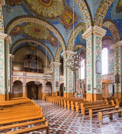 Zdjęcie nr 8: Dekoracja malarska wnętrza kościoła została oparta zasadniczo na motywach ornamentalnych, gdzie wyjątek stanowi monumentalna scena ukazująca uczniów idących do Emaus, usytuowana na północnej ścianie prezbiterium, nad wejściem do zakrystii. Wyobrażono tu w malowanym, ornamentalnym obramieniu scenę wpisaną w stojący prostokąt, zamknięty od góry łukiem półkolistym. W centrum kompozycji ukazano Chrystusa, którego zatrzymują stojący na schodach gospody dwaj uczniowie idący do Emaus. Stojący Zbawiciel został wyobrażony z lewej strony w trzech czwartych w prawo. Prawą ręką podtrzymuje połę płaszcza, lewa jest niewidoczna. Jest to dojrzały mężczyzna o pociągłej twarzy z wydatnym nosem i ustami, długimi włosami spływającymi na ramiona oraz brodą okalającą twarz o jasnej karnacji. Zbawiciel ubrany jest w białą tunikę i szkarłatny płaszcz. Do niego zwraca się jedne z uczniów ukazany tyłem do widza w trzech czwartych w lewo. Jest to dojrzały mężczyzna, który lewą rękę wyciąga w stronę Jezusa, prawą zaś zdaje się zapraszać do wejścia na schody. Jego twarz okolona krótkimi włosami z brodą i wydatnym nosem i ustami widoczna jest z profilu. Ubrany jest w jasną tunikę oraz jasnobeżowy płaszcz. Przed nim z prawej strony na stopniach wiodących do drzwi gospody stoi drugi uczeń. Ujęto go frontalnie w kontrapoście z głową w trzech czwartych w lewo. W prawej ręce trzyma laskę, lewą zaś wskazuje na drzwi zajazdu. Jest to młody mężczyzna o pociągłej twarzy i długich włosach spływających na plecy. Ubrany jest w przewiązaną w pasie tunikę oraz narzucony na ramiona bladoszkarłatny płaszcz. Za przedstawionymi postaciami widać fragment wschodniej architektury oraz rozbudowaną sekwencję pejzażową z elementem bramy miejskiej oraz roślinnością na tle wieczornego nieba. Poniżej przedstawienia umieszczono napis: "Uczniowie z Emaus" podpis: "ZOSTAŃ Z NAMI, BOĆ SIĘ MA KU WIECZOROWI I DZIEŃ SIĘ JUŻ NACHYLIŁ - ŚW[IĘTY] ŁUKASZ XXIV". Bogactwo ornamentalnej dekoracji malarskiej zostało podporządkowane architekturze wnętrza. Partię ścian pokryto malaturą o charakterze dywanowym, ułożoną strefowo z powtarzających się rytmicznie kolorowych motywów ornamentalnych. Gurty sklepienne oraz trzony pilastrów na filarach międzynawowych pokryto wykonanymi na niebieskim lub błękitnym tle ornamentalnymi pasami z motywem fantazyjnych kwiatów i wici roślinnych. Partię sklepienne w kolejnych przęsłach w nawach udekorowano koncentrycznie rozchodzą się płomienistą i falującą ornamentyką, z wplecionymi motywami symbolu Najświętszego Serca Pana Jezusa, Apokaliptycznego Baranka, kielicha z hostią w otoczeniu kłosów zbóż i winnej latorośli, a także gałązek różanych i cierniowych. Na ścianach prezbiterium zastosowano podział strefowy. Dolne przestrzenie ozdobiono pasma ornamentalnymi. Powyżej rozmieszczono strefę z przedstawieniem rosnących obok siebie kwitów lilii. U samej góry sięgnięto do motywów rybich łusek oraz stylizowanej, wielobarwnej ornamentyki fantazyjnej. Gurty sklepienia udekorowano pasami opartymi na stylizowanych skrzydłach serafinów. Natomiast w poszczególnych polach sklepiennych na niebieskim tle w fantazyjnych, falujących otokach wyobrażono dwa skrzyżowane płonące miecze, oko Opatrzności Bożej, dwie tablice Dekalogu. Na środkowej gurcie u szczytu w barwnym obramienium ukazano napis: "KTÓŻ JAK BÓG". 