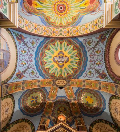 Zdjęcie nr 2: Dekoracja malarska wnętrza kościoła została oparta zasadniczo na motywach ornamentalnych, gdzie wyjątek stanowi monumentalna scena ukazująca uczniów idących do Emaus, usytuowana na północnej ścianie prezbiterium, nad wejściem do zakrystii. Wyobrażono tu w malowanym, ornamentalnym obramieniu scenę wpisaną w stojący prostokąt, zamknięty od góry łukiem półkolistym. W centrum kompozycji ukazano Chrystusa, którego zatrzymują stojący na schodach gospody dwaj uczniowie idący do Emaus. Stojący Zbawiciel został wyobrażony z lewej strony w trzech czwartych w prawo. Prawą ręką podtrzymuje połę płaszcza, lewa jest niewidoczna. Jest to dojrzały mężczyzna o pociągłej twarzy z wydatnym nosem i ustami, długimi włosami spływającymi na ramiona oraz brodą okalającą twarz o jasnej karnacji. Zbawiciel ubrany jest w białą tunikę i szkarłatny płaszcz. Do niego zwraca się jedne z uczniów ukazany tyłem do widza w trzech czwartych w lewo. Jest to dojrzały mężczyzna, który lewą rękę wyciąga w stronę Jezusa, prawą zaś zdaje się zapraszać do wejścia na schody. Jego twarz okolona krótkimi włosami z brodą i wydatnym nosem i ustami widoczna jest z profilu. Ubrany jest w jasną tunikę oraz jasnobeżowy płaszcz. Przed nim z prawej strony na stopniach wiodących do drzwi gospody stoi drugi uczeń. Ujęto go frontalnie w kontrapoście z głową w trzech czwartych w lewo. W prawej ręce trzyma laskę, lewą zaś wskazuje na drzwi zajazdu. Jest to młody mężczyzna o pociągłej twarzy i długich włosach spływających na plecy. Ubrany jest w przewiązaną w pasie tunikę oraz narzucony na ramiona bladoszkarłatny płaszcz. Za przedstawionymi postaciami widać fragment wschodniej architektury oraz rozbudowaną sekwencję pejzażową z elementem bramy miejskiej oraz roślinnością na tle wieczornego nieba. Poniżej przedstawienia umieszczono napis: &quot;Uczniowie z Emaus&quot; podpis: &quot;ZOSTAŃ Z NAMI, BOĆ SIĘ MA KU WIECZOROWI I DZIEŃ SIĘ JUŻ NACHYLIŁ - ŚW[IĘTY] ŁUKASZ XXIV&quot;. Bogactwo ornamentalnej dekoracji malarskiej zostało podporządkowane architekturze wnętrza. Partię ścian pokryto malaturą o charakterze dywanowym, ułożoną strefowo z powtarzających się rytmicznie kolorowych motywów ornamentalnych. Gurty sklepienne oraz trzony pilastrów na filarach międzynawowych pokryto wykonanymi na niebieskim lub błękitnym tle ornamentalnymi pasami z motywem fantazyjnych kwiatów i wici roślinnych. Partię sklepienne w kolejnych przęsłach w nawach udekorowano koncentrycznie rozchodzą się płomienistą i falującą ornamentyką, z wplecionymi motywami symbolu Najświętszego Serca Pana Jezusa, Apokaliptycznego Baranka, kielicha z hostią w otoczeniu kłosów zbóż i winnej latorośli, a także gałązek różanych i cierniowych. Na ścianach prezbiterium zastosowano podział strefowy. Dolne przestrzenie ozdobiono pasma ornamentalnymi. Powyżej rozmieszczono strefę z przedstawieniem rosnących obok siebie kwitów lilii. U samej góry sięgnięto do motywów rybich łusek oraz stylizowanej, wielobarwnej ornamentyki fantazyjnej. Gurty sklepienia udekorowano pasami opartymi na stylizowanych skrzydłach serafinów. Natomiast w poszczególnych polach sklepiennych na niebieskim tle w fantazyjnych, falujących otokach wyobrażono dwa skrzyżowane płonące miecze, oko Opatrzności Bożej, dwie tablice Dekalogu. Na środkowej gurcie u szczytu w barwnym obramienium ukazano napis: &quot;KTÓŻ JAK BÓG&quot;. 