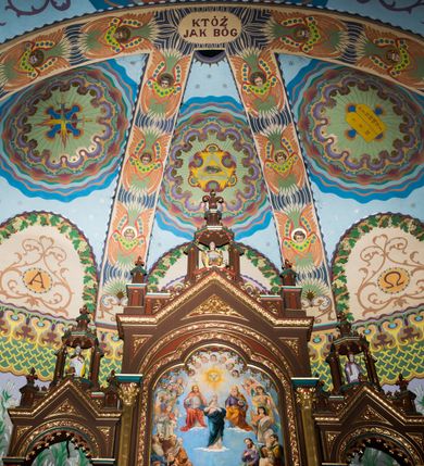 Zdjęcie nr 4: Dekoracja malarska wnętrza kościoła została oparta zasadniczo na motywach ornamentalnych, gdzie wyjątek stanowi monumentalna scena ukazująca uczniów idących do Emaus, usytuowana na północnej ścianie prezbiterium, nad wejściem do zakrystii. Wyobrażono tu w malowanym, ornamentalnym obramieniu scenę wpisaną w stojący prostokąt, zamknięty od góry łukiem półkolistym. W centrum kompozycji ukazano Chrystusa, którego zatrzymują stojący na schodach gospody dwaj uczniowie idący do Emaus. Stojący Zbawiciel został wyobrażony z lewej strony w trzech czwartych w prawo. Prawą ręką podtrzymuje połę płaszcza, lewa jest niewidoczna. Jest to dojrzały mężczyzna o pociągłej twarzy z wydatnym nosem i ustami, długimi włosami spływającymi na ramiona oraz brodą okalającą twarz o jasnej karnacji. Zbawiciel ubrany jest w białą tunikę i szkarłatny płaszcz. Do niego zwraca się jedne z uczniów ukazany tyłem do widza w trzech czwartych w lewo. Jest to dojrzały mężczyzna, który lewą rękę wyciąga w stronę Jezusa, prawą zaś zdaje się zapraszać do wejścia na schody. Jego twarz okolona krótkimi włosami z brodą i wydatnym nosem i ustami widoczna jest z profilu. Ubrany jest w jasną tunikę oraz jasnobeżowy płaszcz. Przed nim z prawej strony na stopniach wiodących do drzwi gospody stoi drugi uczeń. Ujęto go frontalnie w kontrapoście z głową w trzech czwartych w lewo. W prawej ręce trzyma laskę, lewą zaś wskazuje na drzwi zajazdu. Jest to młody mężczyzna o pociągłej twarzy i długich włosach spływających na plecy. Ubrany jest w przewiązaną w pasie tunikę oraz narzucony na ramiona bladoszkarłatny płaszcz. Za przedstawionymi postaciami widać fragment wschodniej architektury oraz rozbudowaną sekwencję pejzażową z elementem bramy miejskiej oraz roślinnością na tle wieczornego nieba. Poniżej przedstawienia umieszczono napis: "Uczniowie z Emaus" podpis: "ZOSTAŃ Z NAMI, BOĆ SIĘ MA KU WIECZOROWI I DZIEŃ SIĘ JUŻ NACHYLIŁ - ŚW[IĘTY] ŁUKASZ XXIV". Bogactwo ornamentalnej dekoracji malarskiej zostało podporządkowane architekturze wnętrza. Partię ścian pokryto malaturą o charakterze dywanowym, ułożoną strefowo z powtarzających się rytmicznie kolorowych motywów ornamentalnych. Gurty sklepienne oraz trzony pilastrów na filarach międzynawowych pokryto wykonanymi na niebieskim lub błękitnym tle ornamentalnymi pasami z motywem fantazyjnych kwiatów i wici roślinnych. Partię sklepienne w kolejnych przęsłach w nawach udekorowano koncentrycznie rozchodzą się płomienistą i falującą ornamentyką, z wplecionymi motywami symbolu Najświętszego Serca Pana Jezusa, Apokaliptycznego Baranka, kielicha z hostią w otoczeniu kłosów zbóż i winnej latorośli, a także gałązek różanych i cierniowych. Na ścianach prezbiterium zastosowano podział strefowy. Dolne przestrzenie ozdobiono pasma ornamentalnymi. Powyżej rozmieszczono strefę z przedstawieniem rosnących obok siebie kwitów lilii. U samej góry sięgnięto do motywów rybich łusek oraz stylizowanej, wielobarwnej ornamentyki fantazyjnej. Gurty sklepienia udekorowano pasami opartymi na stylizowanych skrzydłach serafinów. Natomiast w poszczególnych polach sklepiennych na niebieskim tle w fantazyjnych, falujących otokach wyobrażono dwa skrzyżowane płonące miecze, oko Opatrzności Bożej, dwie tablice Dekalogu. Na środkowej gurcie u szczytu w barwnym obramienium ukazano napis: "KTÓŻ JAK BÓG". 