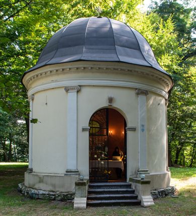 Zdjęcie nr 14: Kaplicę wzniesiono na rzucie koła, nakrywając jej wnętrze półkolistą kopułą. Ściany zewnętrzne zostały podzielone ośmioma pilastrami jońskimi, wspierającymi belkowanie. Powyżej belkowania biegnie profilowany gzyms koronujący, dekorowany fryzem kostkowym. Półokrągłe wejście usytuowano od zachodu. Prowadzi do niego pięć stopni. Wejście zamyka żelazna, klasycystyczna krata. Od wschodu w ścianie przepruto okulus, ujęty od wnętrza półokrągłym glifem w płytkiej, prostokątnej niszy zamkniętej półokrągło. Wnętrze pokryto ochrową polichromią, z wymalowanym białą kreską podziałem odzwierciedlającym artykulację zewnętrzną. Na ścianie umieszczono dwie czworoboczne płyty upamiętniające zmarłych pochowanych w krypcie. Wejście do krypty zamyka prostokątna, marmurowa płyta dekorowana aplikacją ze złoconego brązu, ukazującą trupią czaszkę ze skrzyżowanymi piszczelami umieszczoną nad antabą.