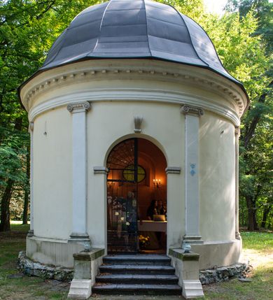 Zdjęcie nr 17: Kaplicę wzniesiono na rzucie koła, nakrywając jej wnętrze półkolistą kopułą. Ściany zewnętrzne zostały podzielone ośmioma pilastrami jońskimi, wspierającymi belkowanie. Powyżej belkowania biegnie profilowany gzyms koronujący, dekorowany fryzem kostkowym. Półokrągłe wejście usytuowano od zachodu. Prowadzi do niego pięć stopni. Wejście zamyka żelazna, klasycystyczna krata. Od wschodu w ścianie przepruto okulus, ujęty od wnętrza półokrągłym glifem w płytkiej, prostokątnej niszy zamkniętej półokrągło. Wnętrze pokryto ochrową polichromią, z wymalowanym białą kreską podziałem odzwierciedlającym artykulację zewnętrzną. Na ścianie umieszczono dwie czworoboczne płyty upamiętniające zmarłych pochowanych w krypcie. Wejście do krypty zamyka prostokątna, marmurowa płyta dekorowana aplikacją ze złoconego brązu, ukazującą trupią czaszkę ze skrzyżowanymi piszczelami umieszczoną nad antabą.