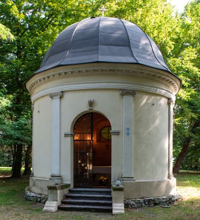 Zdjęcie nr 16: Kaplicę wzniesiono na rzucie koła, nakrywając jej wnętrze półkolistą kopułą. Ściany zewnętrzne zostały podzielone ośmioma pilastrami jońskimi, wspierającymi belkowanie. Powyżej belkowania biegnie profilowany gzyms koronujący, dekorowany fryzem kostkowym. Półokrągłe wejście usytuowano od zachodu. Prowadzi do niego pięć stopni. Wejście zamyka żelazna, klasycystyczna krata. Od wschodu w ścianie przepruto okulus, ujęty od wnętrza półokrągłym glifem w płytkiej, prostokątnej niszy zamkniętej półokrągło. Wnętrze pokryto ochrową polichromią, z wymalowanym białą kreską podziałem odzwierciedlającym artykulację zewnętrzną. Na ścianie umieszczono dwie czworoboczne płyty upamiętniające zmarłych pochowanych w krypcie. Wejście do krypty zamyka prostokątna, marmurowa płyta dekorowana aplikacją ze złoconego brązu, ukazującą trupią czaszkę ze skrzyżowanymi piszczelami umieszczoną nad antabą.