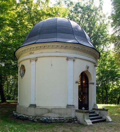 Zdjęcie nr 13: Kaplicę wzniesiono na rzucie koła, nakrywając jej wnętrze półkolistą kopułą. Ściany zewnętrzne zostały podzielone ośmioma pilastrami jońskimi, wspierającymi belkowanie. Powyżej belkowania biegnie profilowany gzyms koronujący, dekorowany fryzem kostkowym. Półokrągłe wejście usytuowano od zachodu. Prowadzi do niego pięć stopni. Wejście zamyka żelazna, klasycystyczna krata. Od wschodu w ścianie przepruto okulus, ujęty od wnętrza półokrągłym glifem w płytkiej, prostokątnej niszy zamkniętej półokrągło. Wnętrze pokryto ochrową polichromią, z wymalowanym białą kreską podziałem odzwierciedlającym artykulację zewnętrzną. Na ścianie umieszczono dwie czworoboczne płyty upamiętniające zmarłych pochowanych w krypcie. Wejście do krypty zamyka prostokątna, marmurowa płyta dekorowana aplikacją ze złoconego brązu, ukazującą trupią czaszkę ze skrzyżowanymi piszczelami umieszczoną nad antabą.