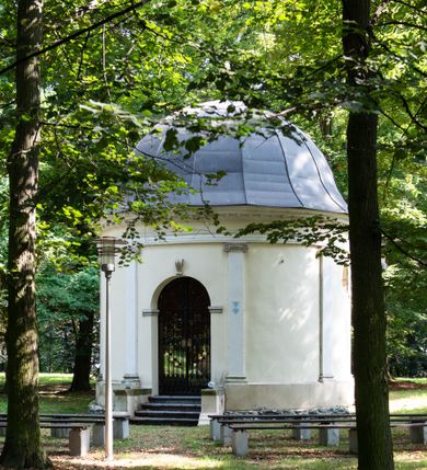 Zdjęcie nr 12: Kaplicę wzniesiono na rzucie koła, nakrywając jej wnętrze półkolistą kopułą. Ściany zewnętrzne zostały podzielone ośmioma pilastrami jońskimi, wspierającymi belkowanie. Powyżej belkowania biegnie profilowany gzyms koronujący, dekorowany fryzem kostkowym. Półokrągłe wejście usytuowano od zachodu. Prowadzi do niego pięć stopni. Wejście zamyka żelazna, klasycystyczna krata. Od wschodu w ścianie przepruto okulus, ujęty od wnętrza półokrągłym glifem w płytkiej, prostokątnej niszy zamkniętej półokrągło. Wnętrze pokryto ochrową polichromią, z wymalowanym białą kreską podziałem odzwierciedlającym artykulację zewnętrzną. Na ścianie umieszczono dwie czworoboczne płyty upamiętniające zmarłych pochowanych w krypcie. Wejście do krypty zamyka prostokątna, marmurowa płyta dekorowana aplikacją ze złoconego brązu, ukazującą trupią czaszkę ze skrzyżowanymi piszczelami umieszczoną nad antabą.