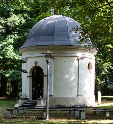 Zdjęcie nr 11: Kaplicę wzniesiono na rzucie koła, nakrywając jej wnętrze półkolistą kopułą. Ściany zewnętrzne zostały podzielone ośmioma pilastrami jońskimi, wspierającymi belkowanie. Powyżej belkowania biegnie profilowany gzyms koronujący, dekorowany fryzem kostkowym. Półokrągłe wejście usytuowano od zachodu. Prowadzi do niego pięć stopni. Wejście zamyka żelazna, klasycystyczna krata. Od wschodu w ścianie przepruto okulus, ujęty od wnętrza półokrągłym glifem w płytkiej, prostokątnej niszy zamkniętej półokrągło. Wnętrze pokryto ochrową polichromią, z wymalowanym białą kreską podziałem odzwierciedlającym artykulację zewnętrzną. Na ścianie umieszczono dwie czworoboczne płyty upamiętniające zmarłych pochowanych w krypcie. Wejście do krypty zamyka prostokątna, marmurowa płyta dekorowana aplikacją ze złoconego brązu, ukazującą trupią czaszkę ze skrzyżowanymi piszczelami umieszczoną nad antabą.